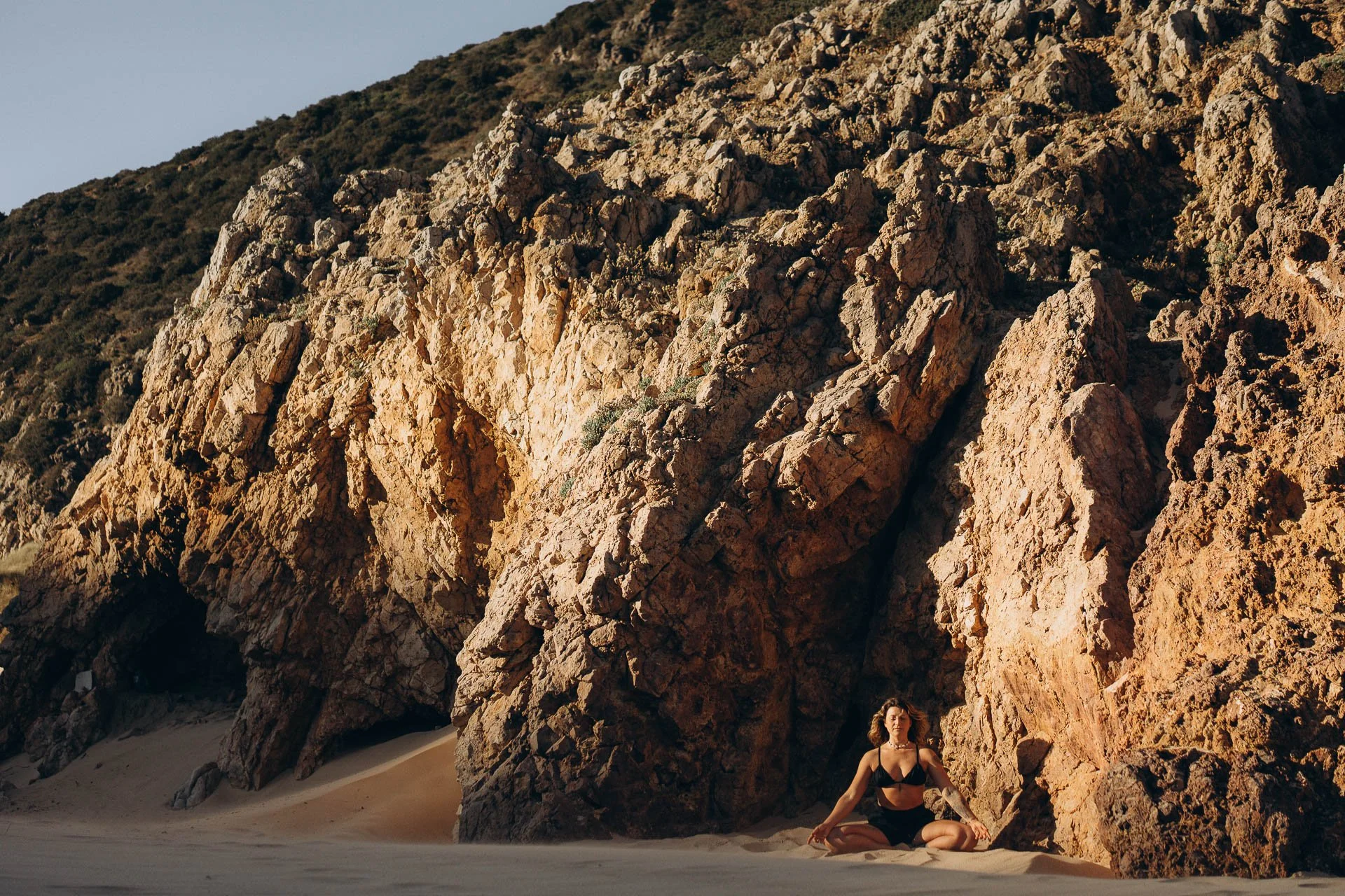 Seated meditation on the beach, surrounded by rock formations and soft reflected light.