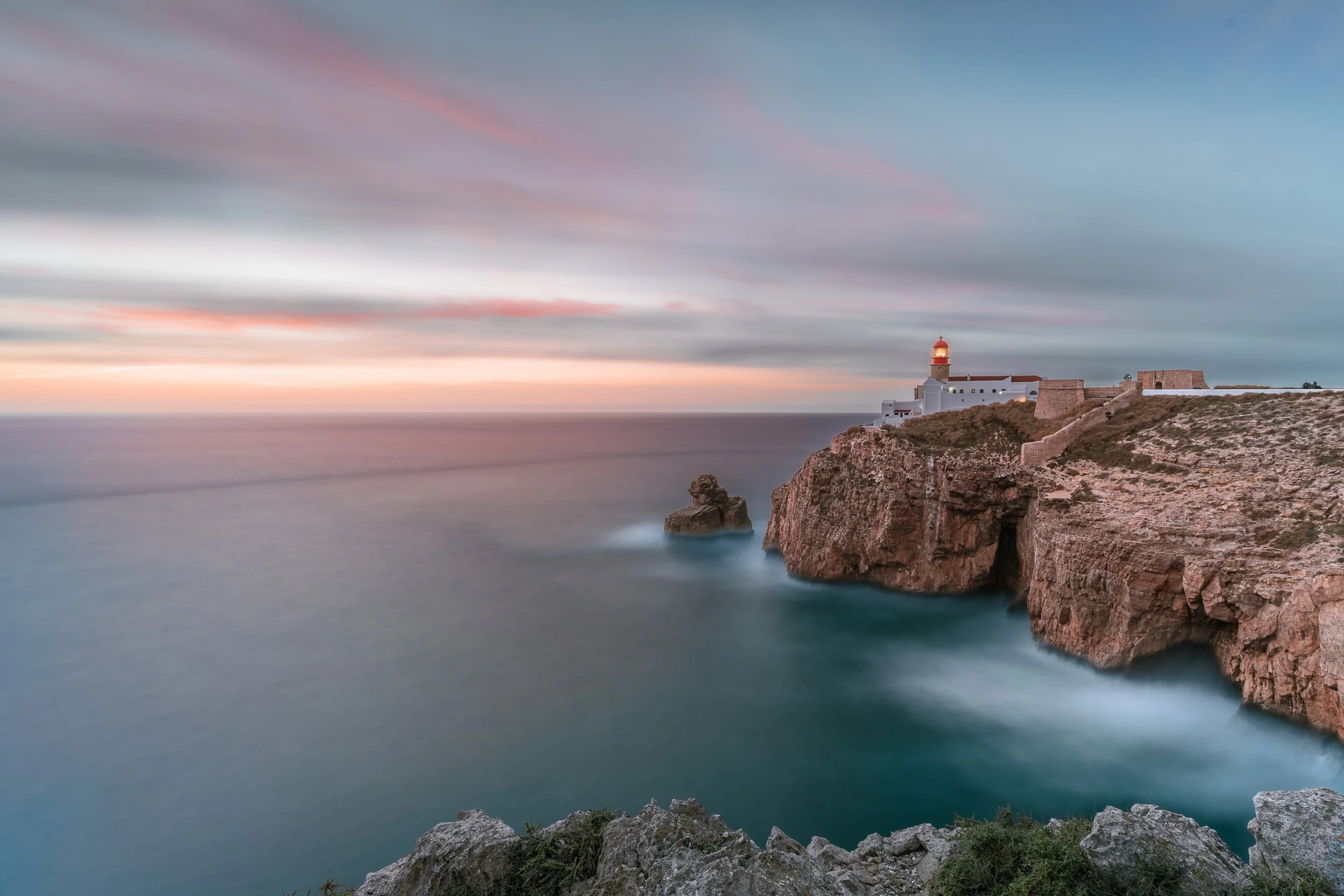Cliffside lighthouse at sunset with a cloudy sky and calm ocean, rocky coastline in the foreground.