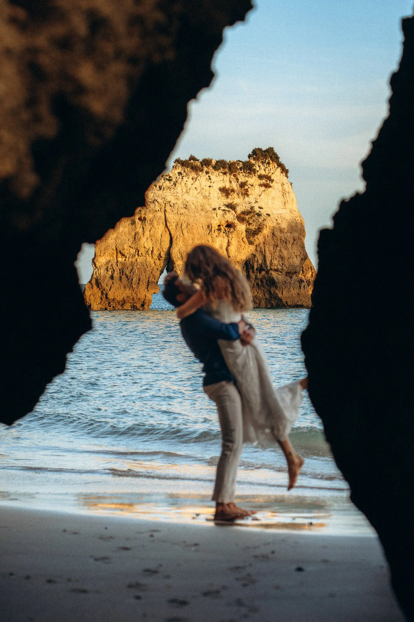 Couple embracing through Algarve sea cave frame, iconic rock arch and ocean behind, engagement photography Lagos