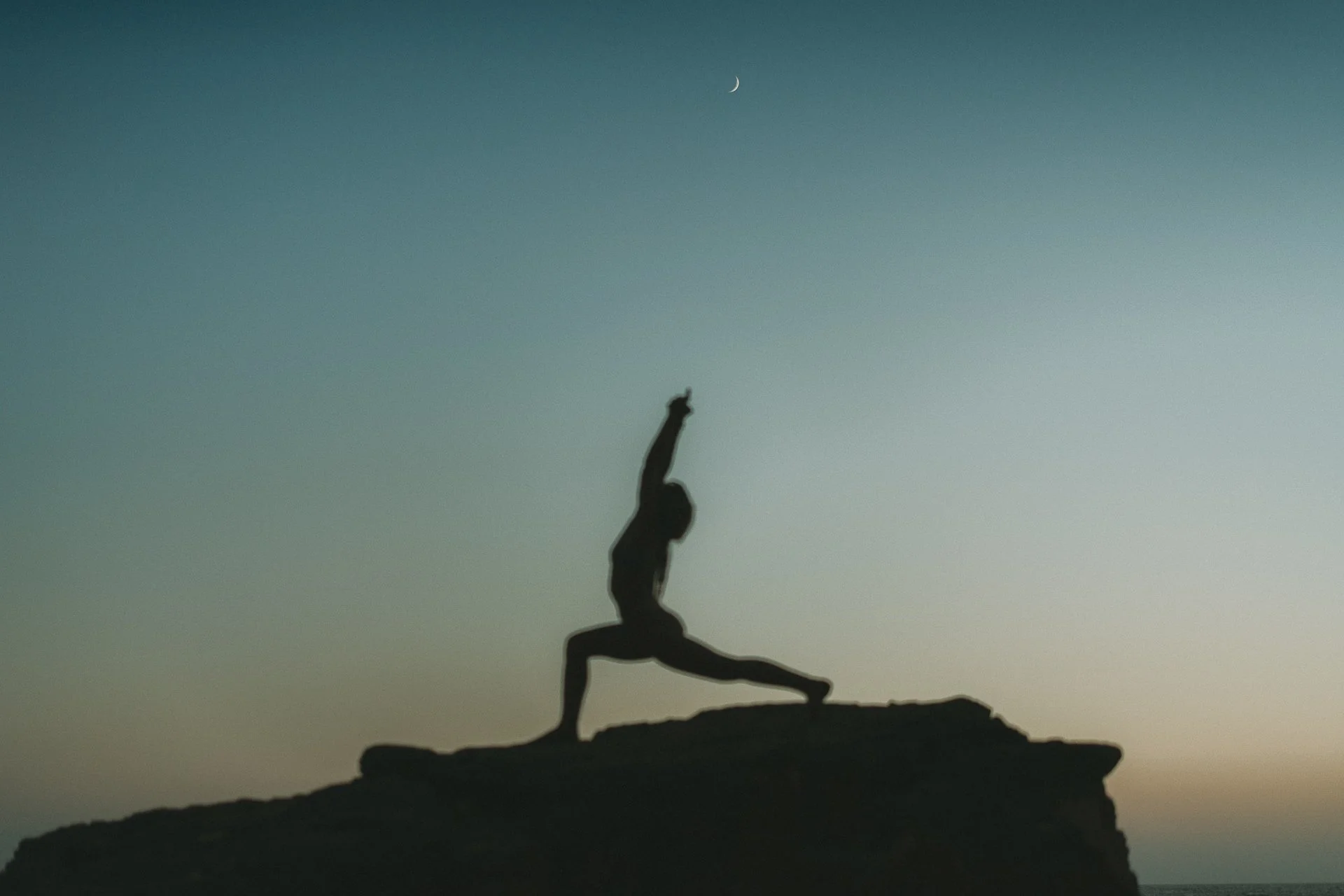 Silhouette yoga pose at dusk, strong warrior stance against a pastel sky and crescent moon.