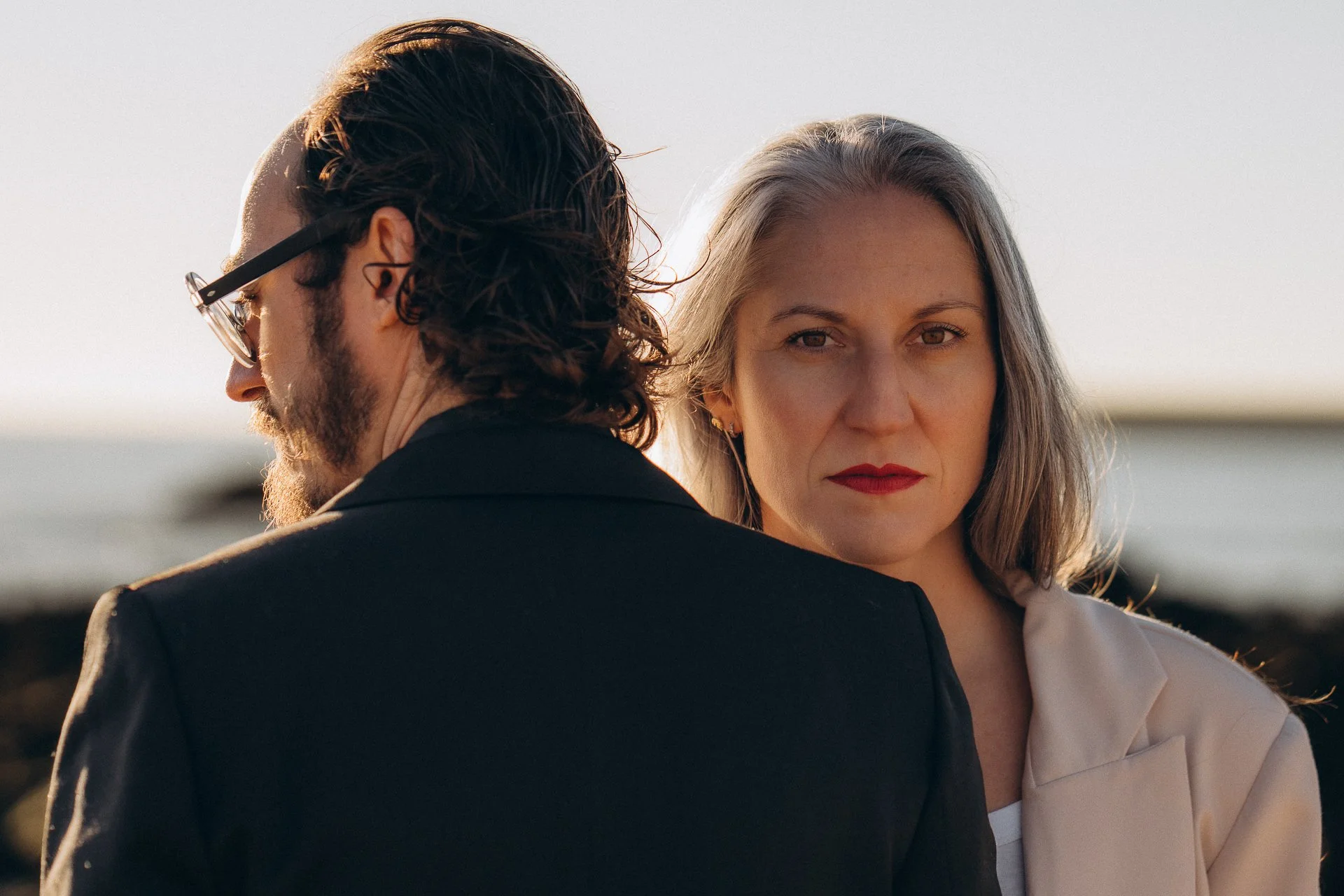 A man with long hair, glasses, and a beard with his back to the camera, and a woman with shoulder-length hair and red lipstick looking directly at the camera, outdoors during sunset.