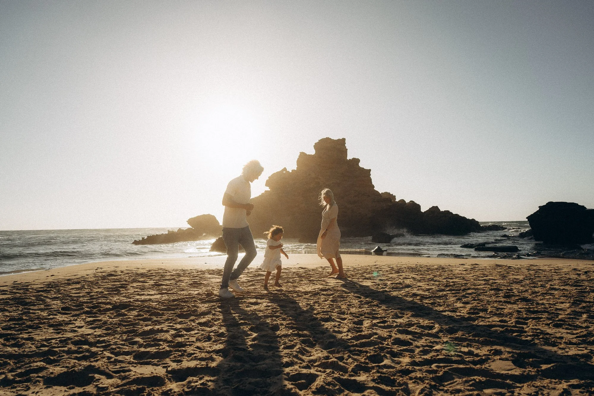 Silhouette of family of three walking on Algarve beach at sunset, dramatic rock formation and sunflare