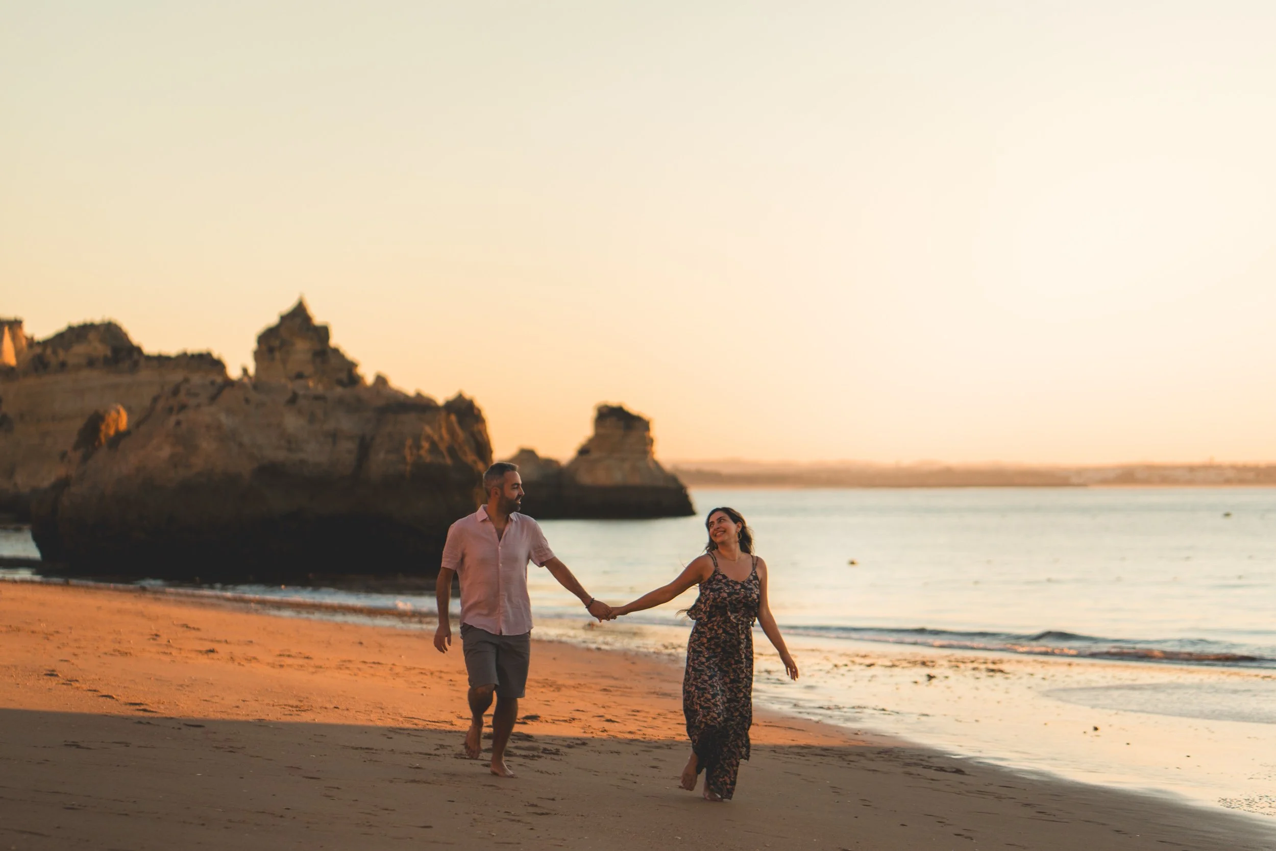 Romantic couple running barefoot on Praia Dona Ana at sunset.