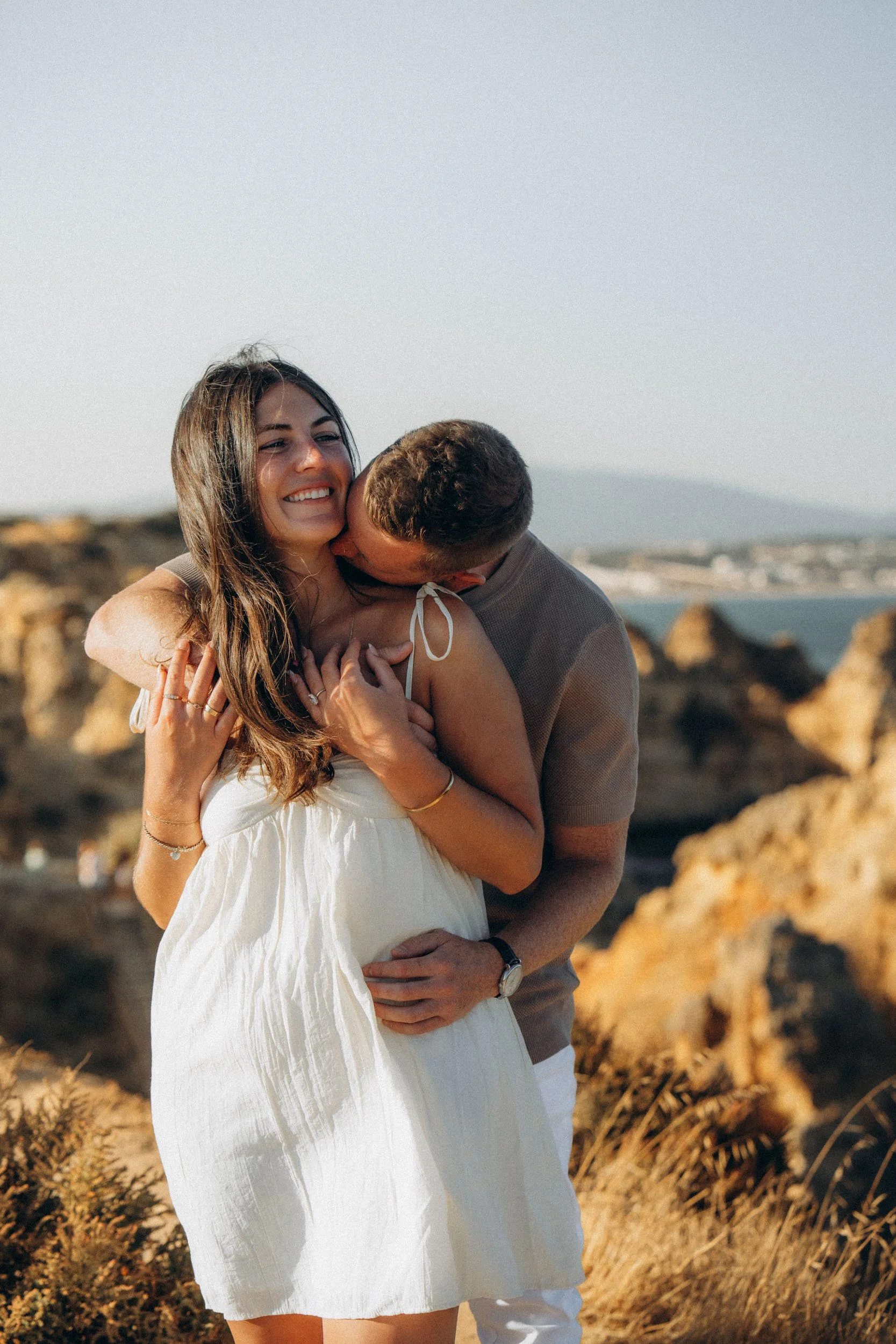 Playful post-proposal moment with ocean breeze and sunset light. Genuine smiles, real reactions.