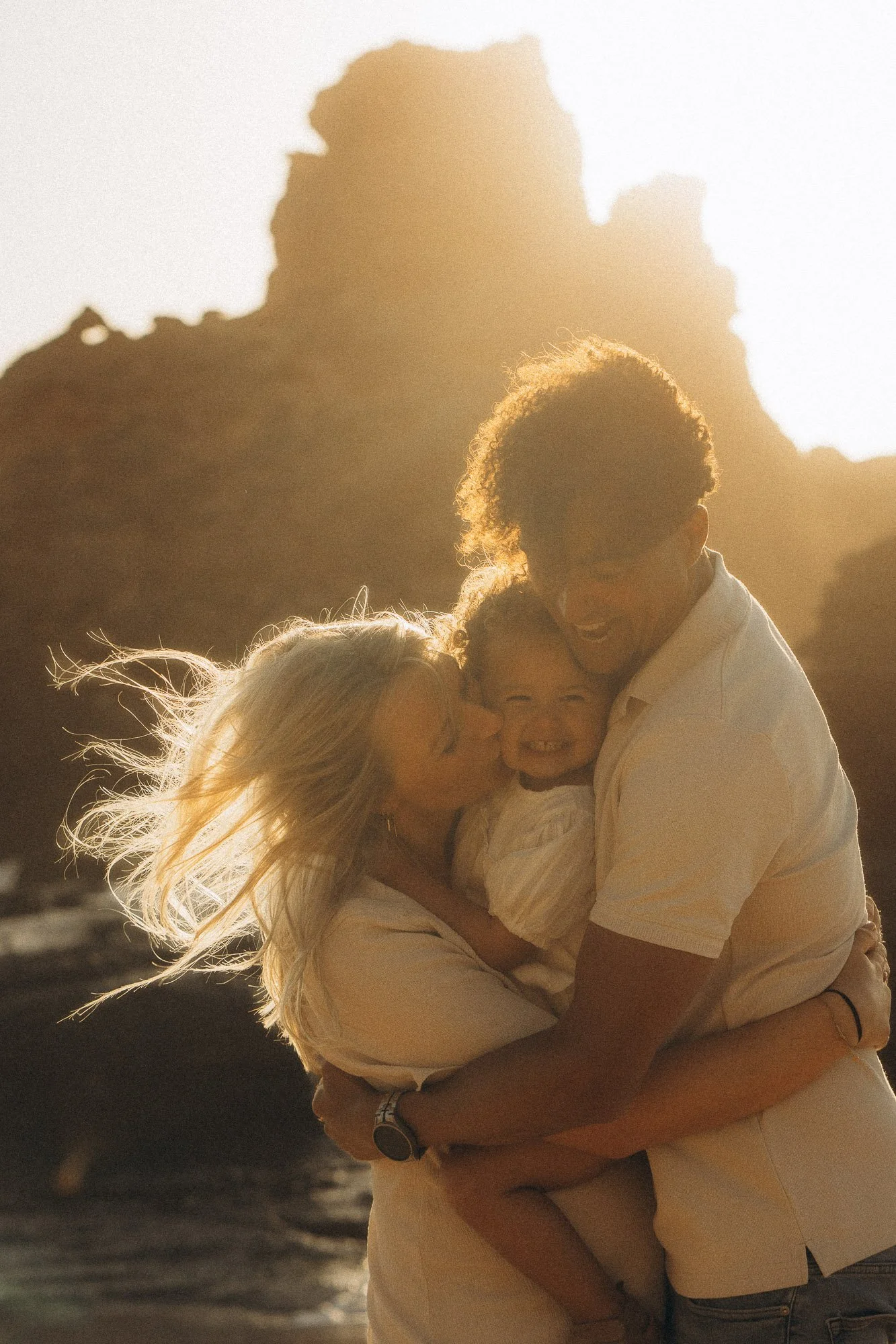 Family photographer Algarve capturing parents and toddler in a warm embrace at sunset, golden backlight and rocky coastline