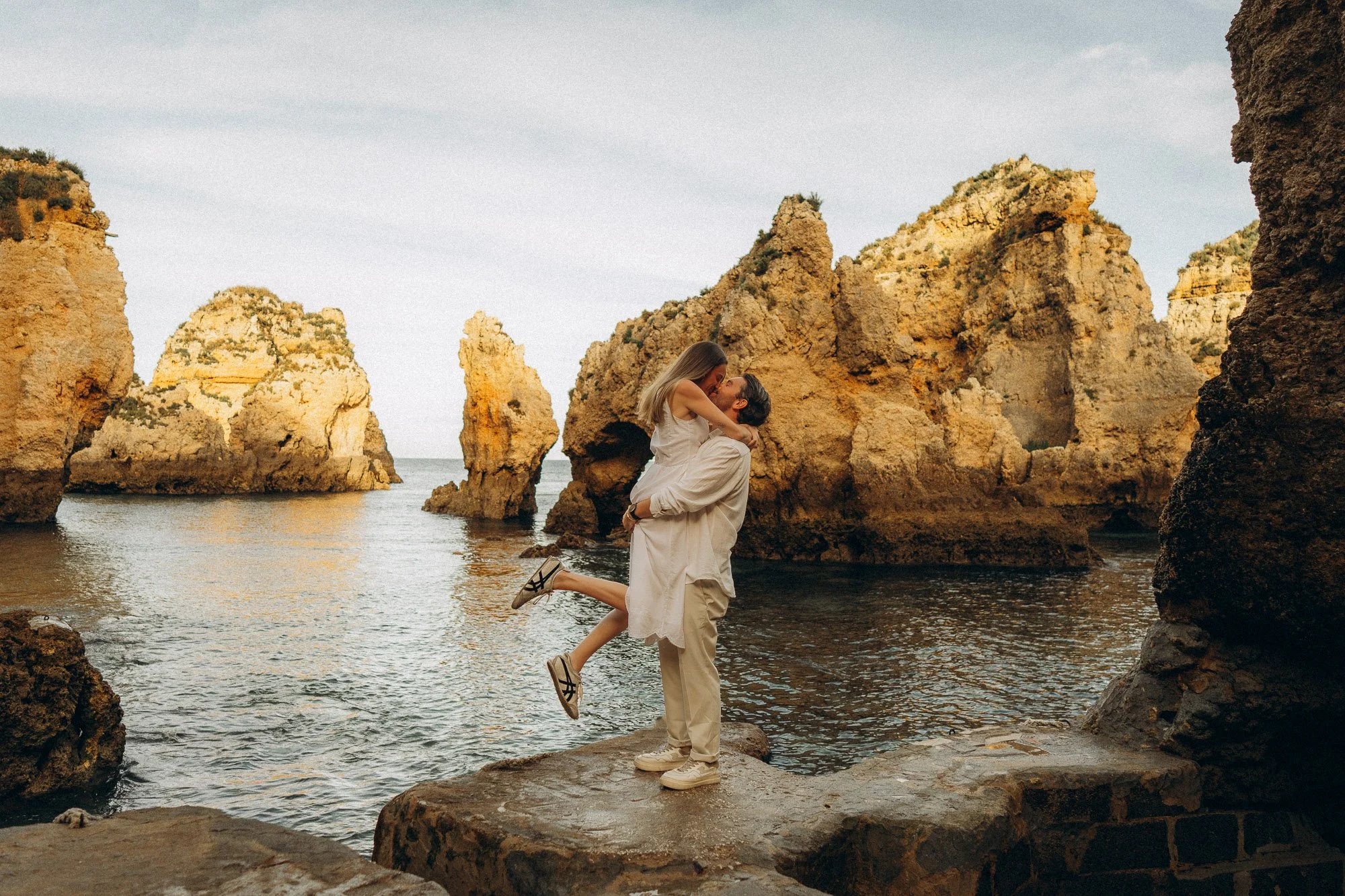 Couple kissing at Ponta da Piedade Lagos, man lifting woman on rocks with sea stacks behind, engagement photographer Algarve