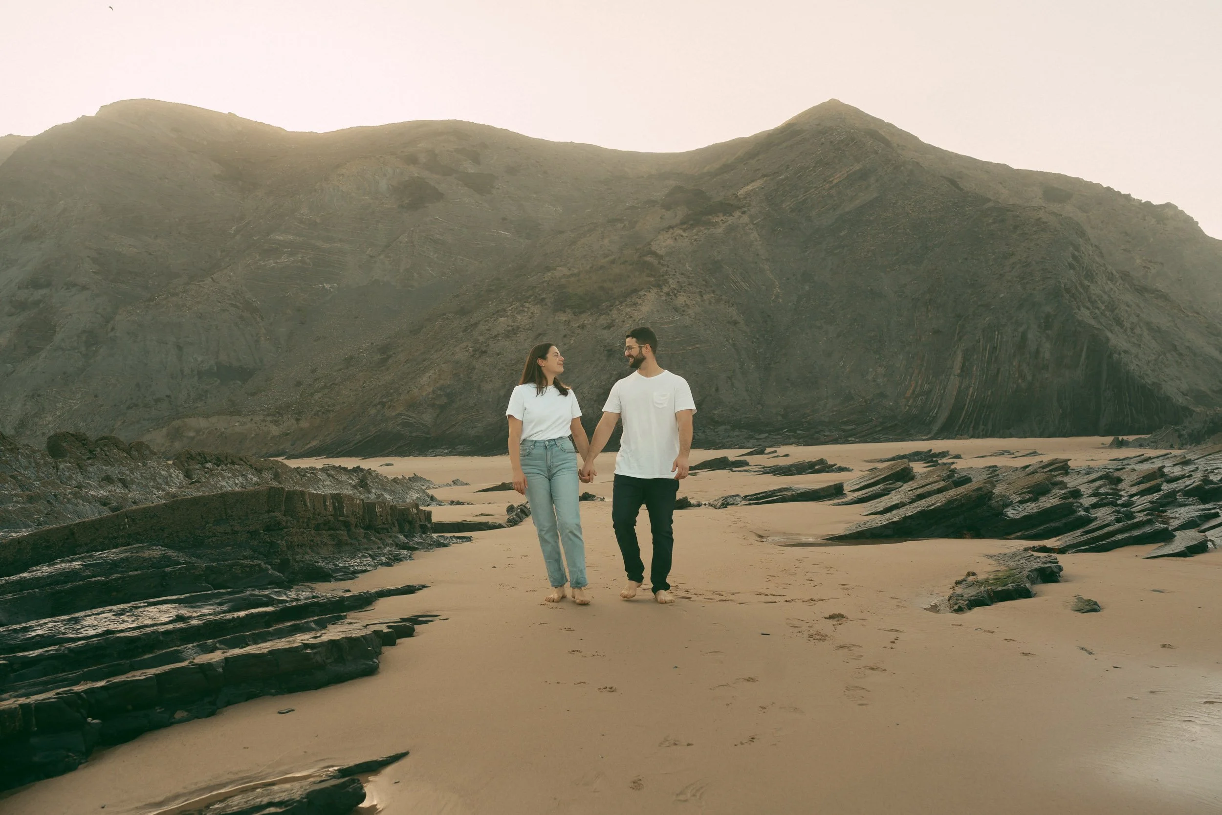 A couple holding hands walking through shallow shoreline water at Praia do Castelejo. Documentary-style couple photoshoot in the Algarve, capturing candid movement and relationship storytelling.