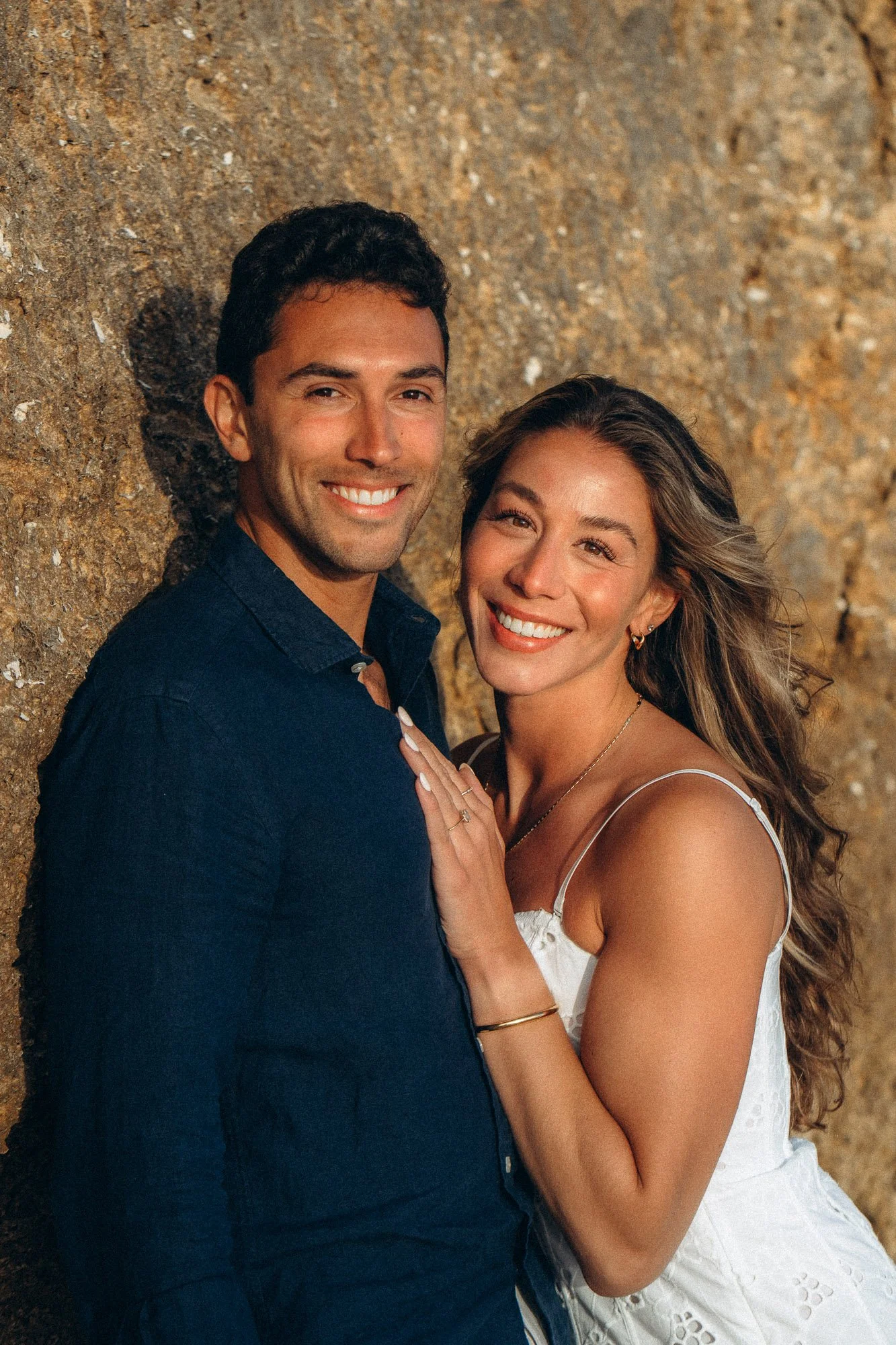 Engaged couple portrait against golden rock wall in Algarve, woman showing engagement ring, natural light