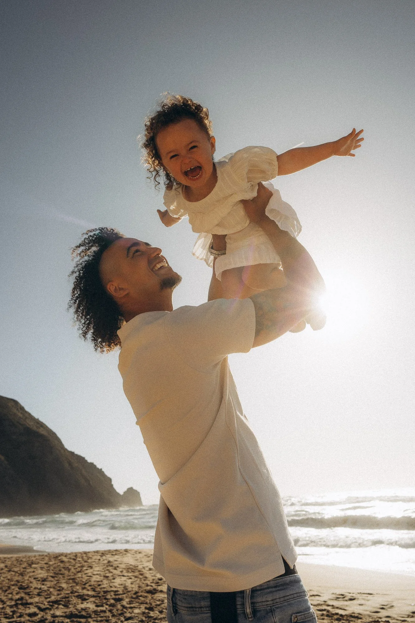 Father lifting laughing toddler above his head during family photography session in Algarve, backlit by sun