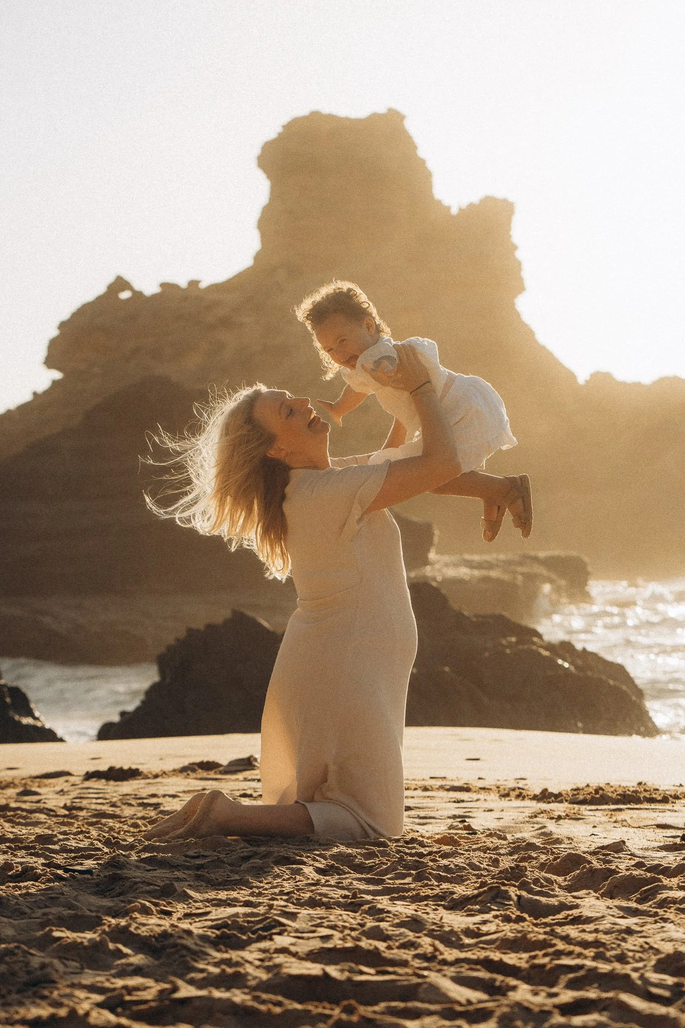 Mother lifting baby up during family photo session on Algarve beach, golden hour light and coastal cliffs