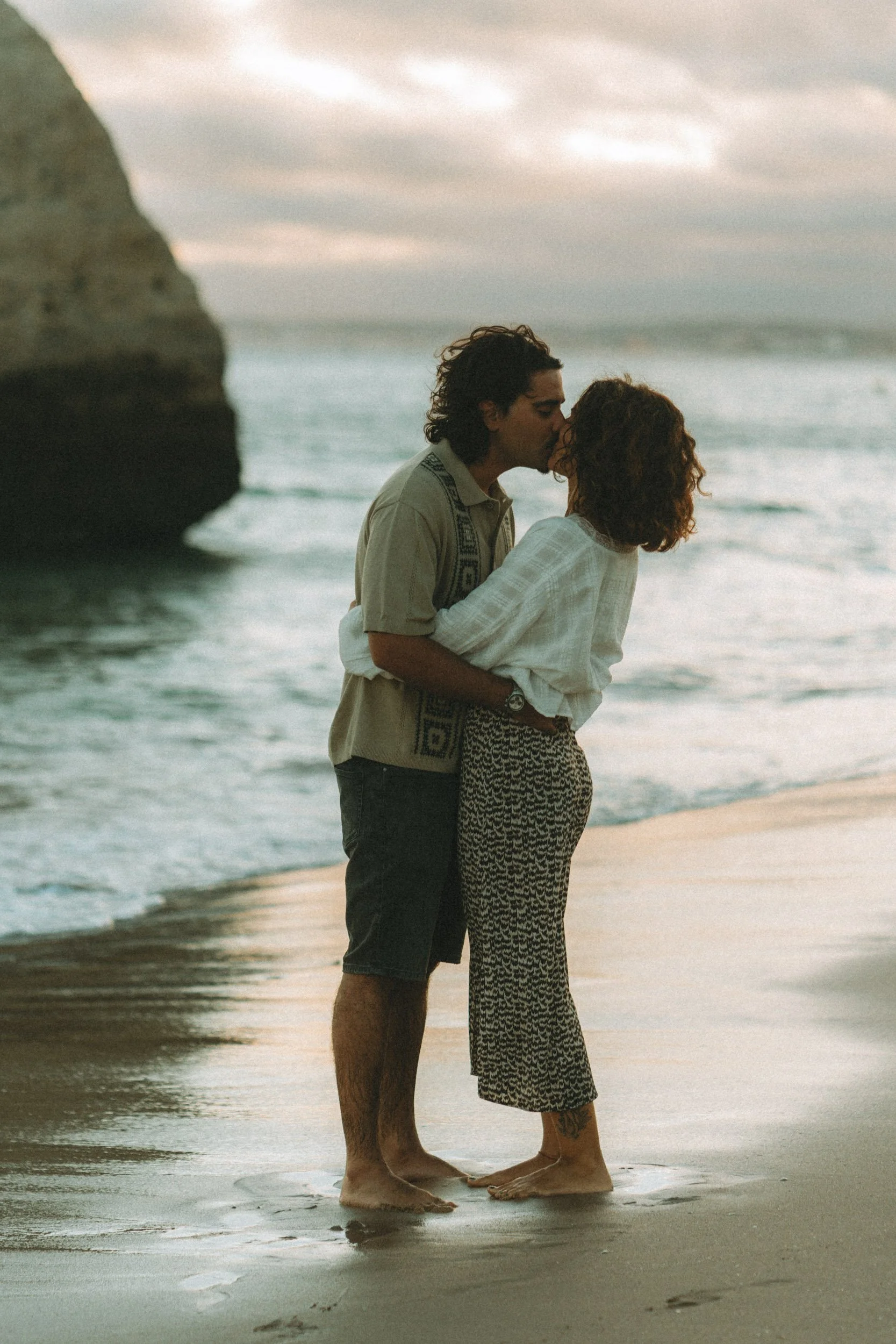 Silhouette-style kiss at sunset on Praia dos Três Irmãos, warm sky and textured ocean creating a timeless romantic mood.