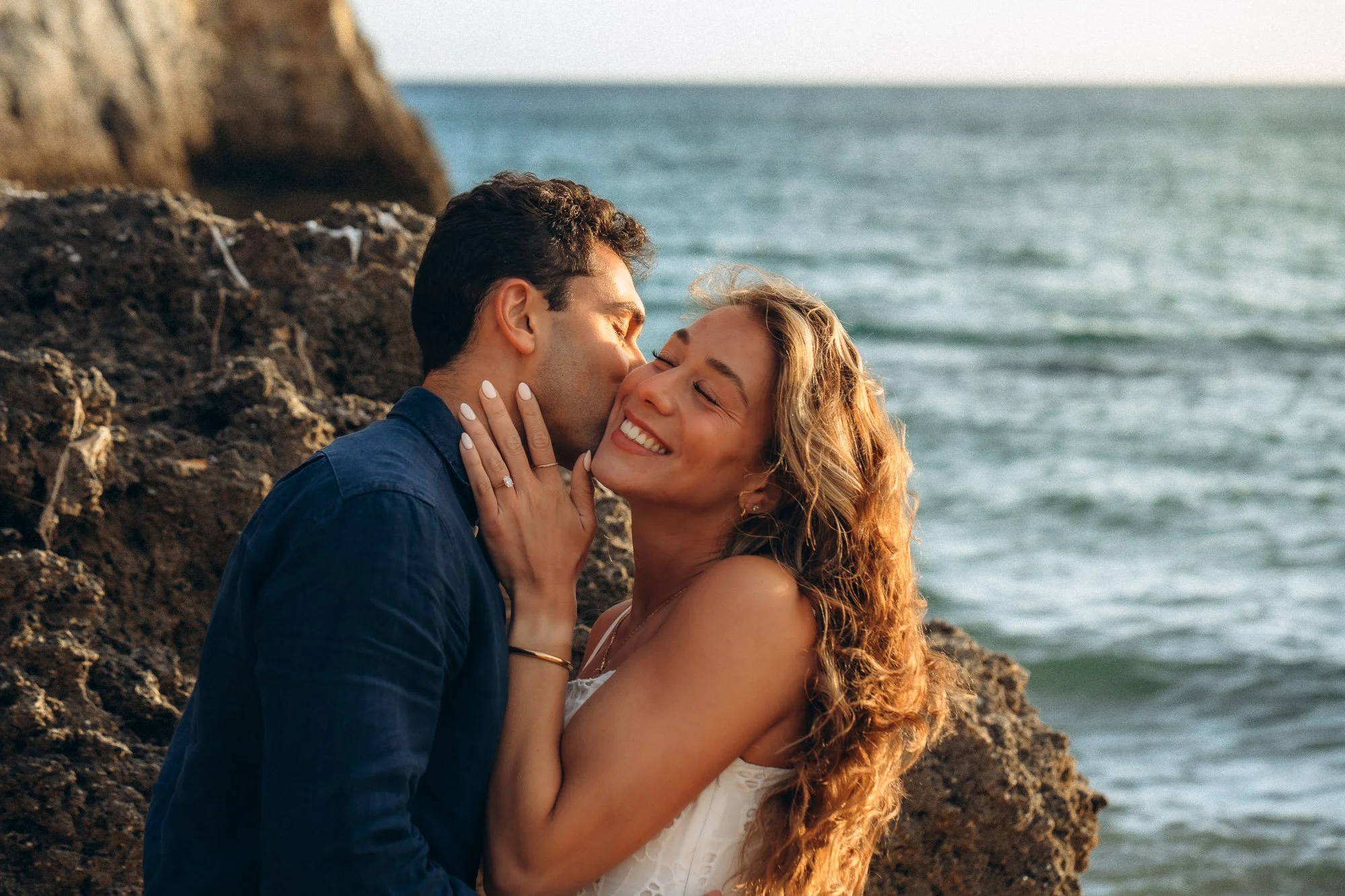 Newly engaged couple kissing on Algarve coastal rocks at golden hour, engagement ring detail
