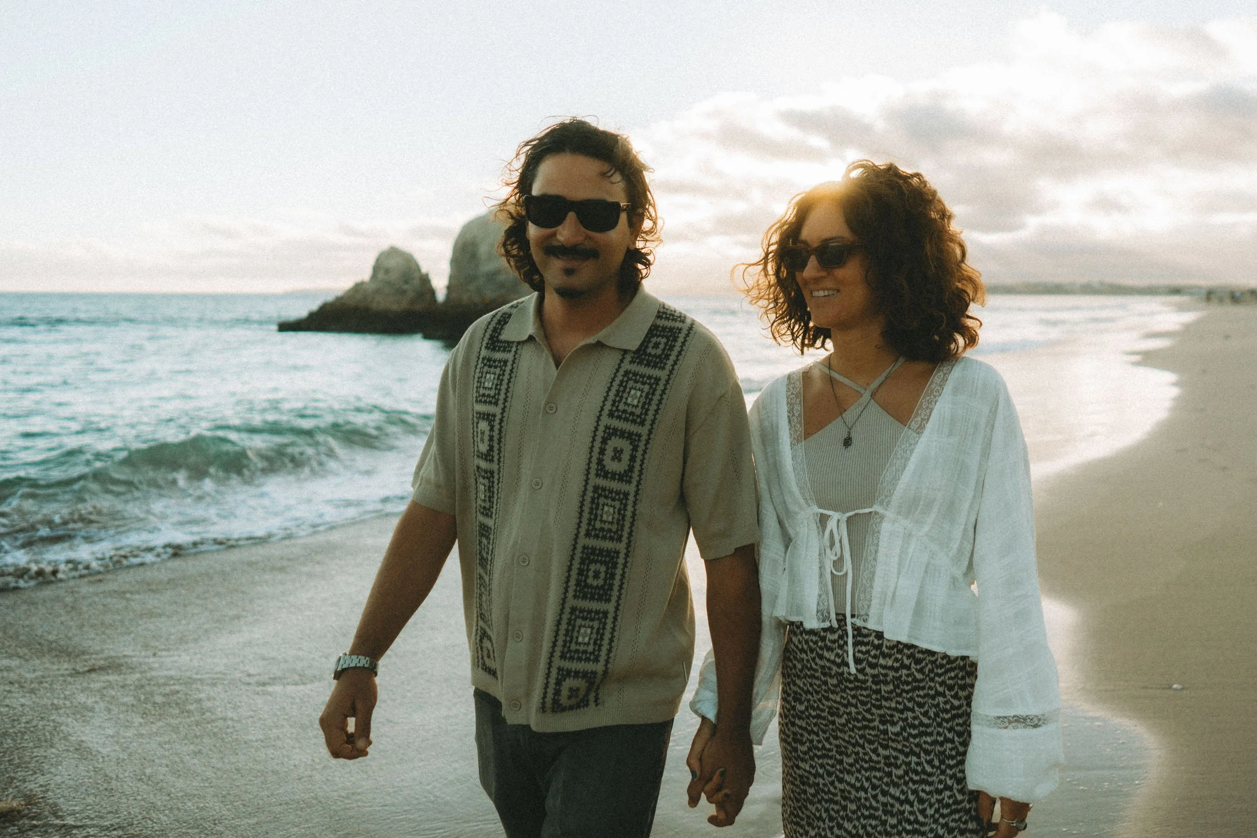 Couple walking hand in hand at Praia dos Três Irmãos during golden hour, with iconic rock formations in the background and soft sunset light reflecting on the water.