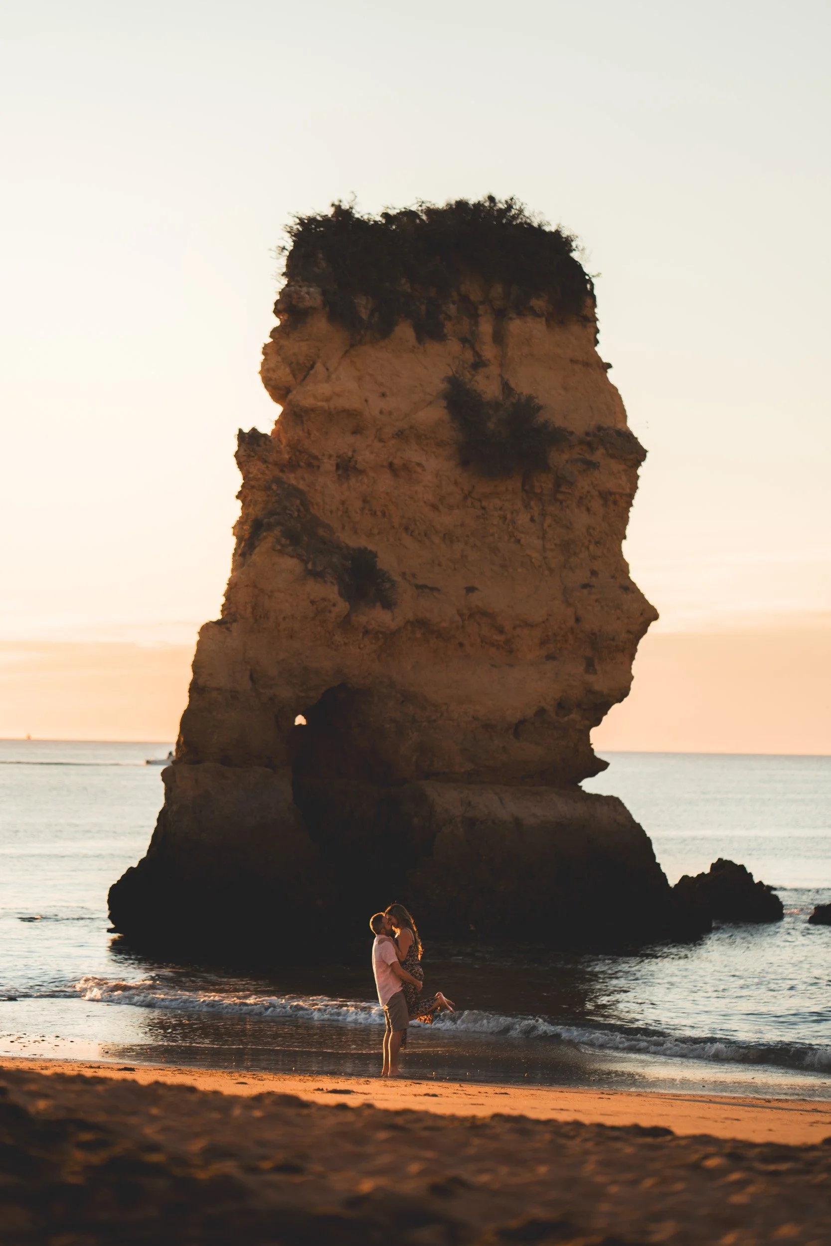 A couple sharing a kiss beneath the iconic rock formation of Praia Dona Ana, Algarve sunset creating warm reflections.