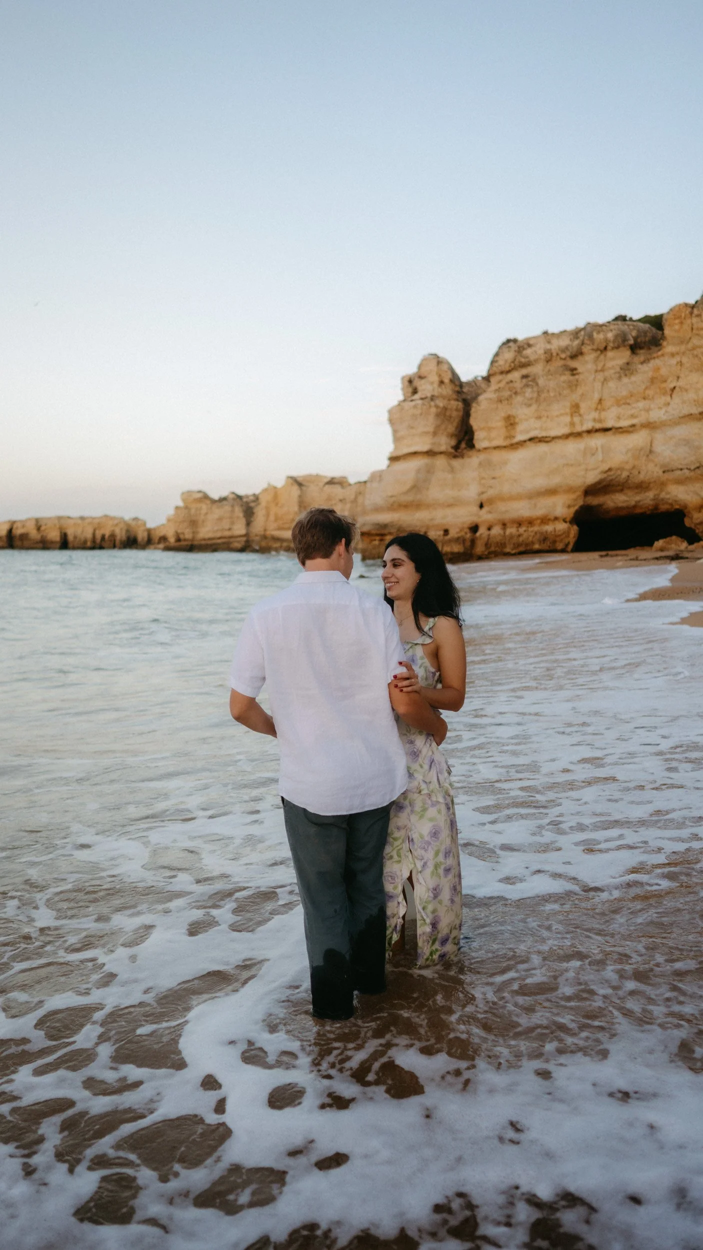 Standing in the ocean as waves gently surround them. A romantic, natural moment with Algarve cliffs glowing in the background.