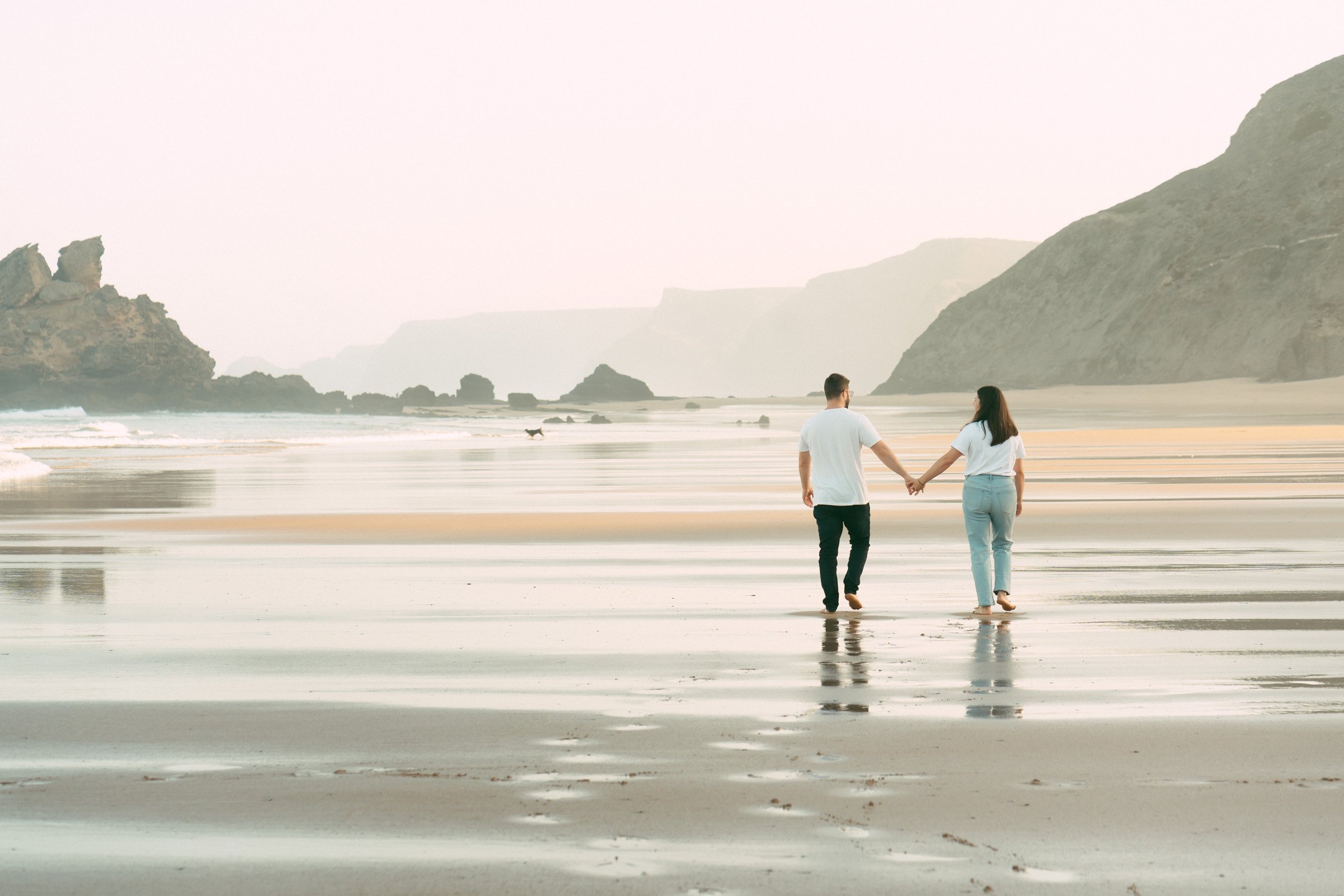 A couple walking hand in hand along wet sand reflections at Praia do Castelejo, Algarve. Candid destination couple photography session capturing connection and movement on the Atlantic coast.