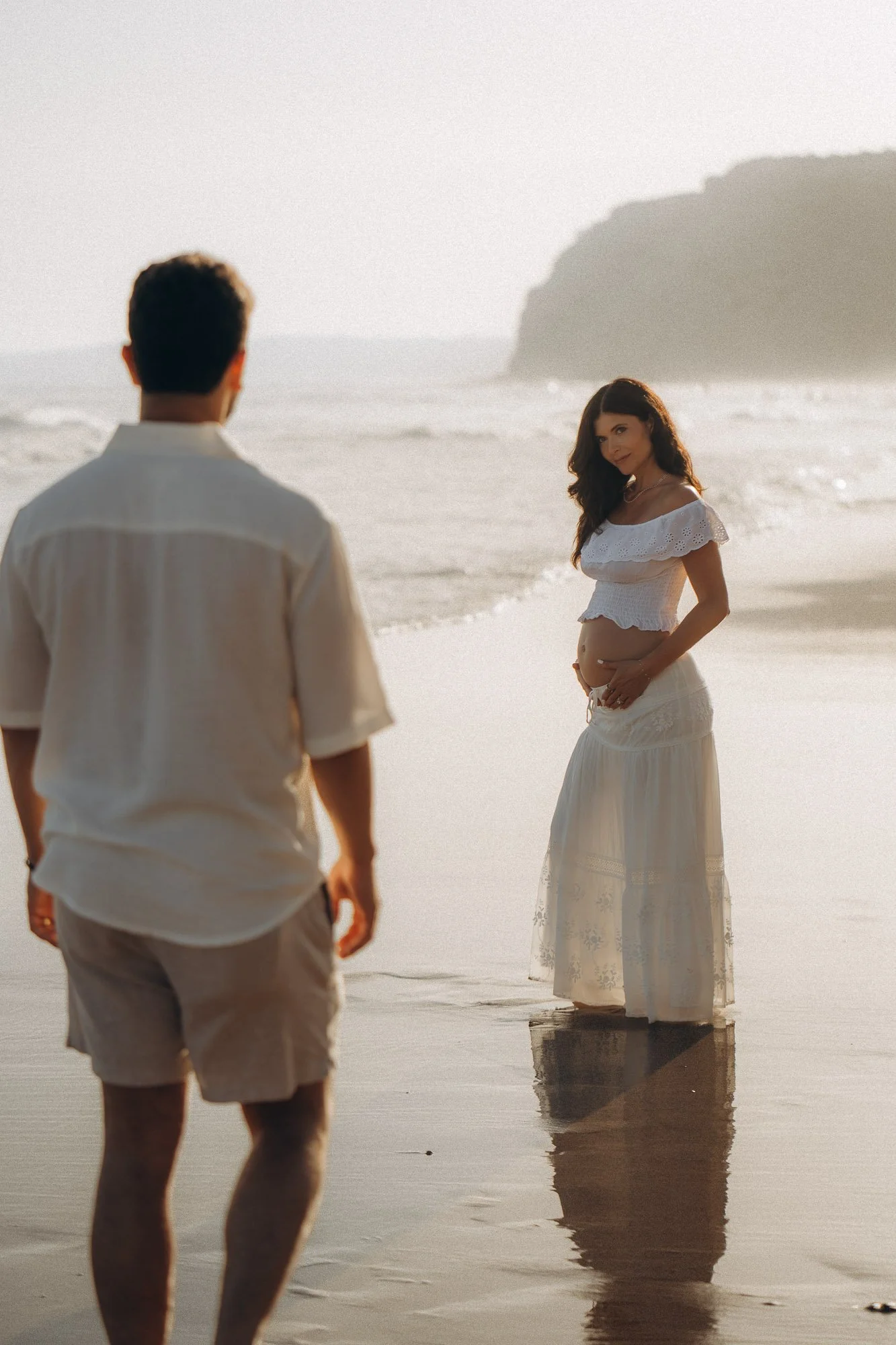 Pregnant woman looking back at partner on misty Algarve beach, cinematic maternity photography Portugal