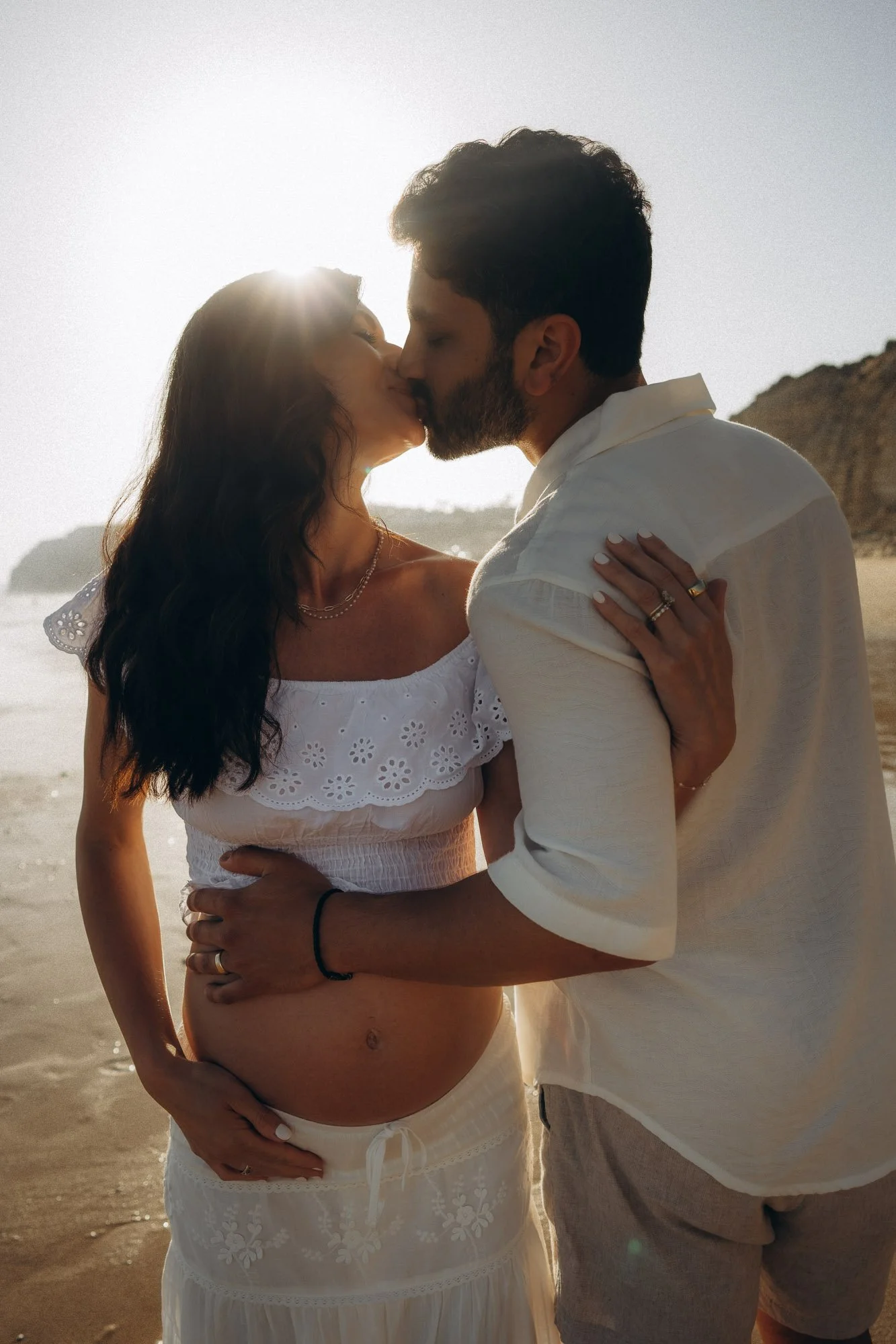 Maternity photography Algarve, couple kissing with baby bump, backlit golden hour on beach
