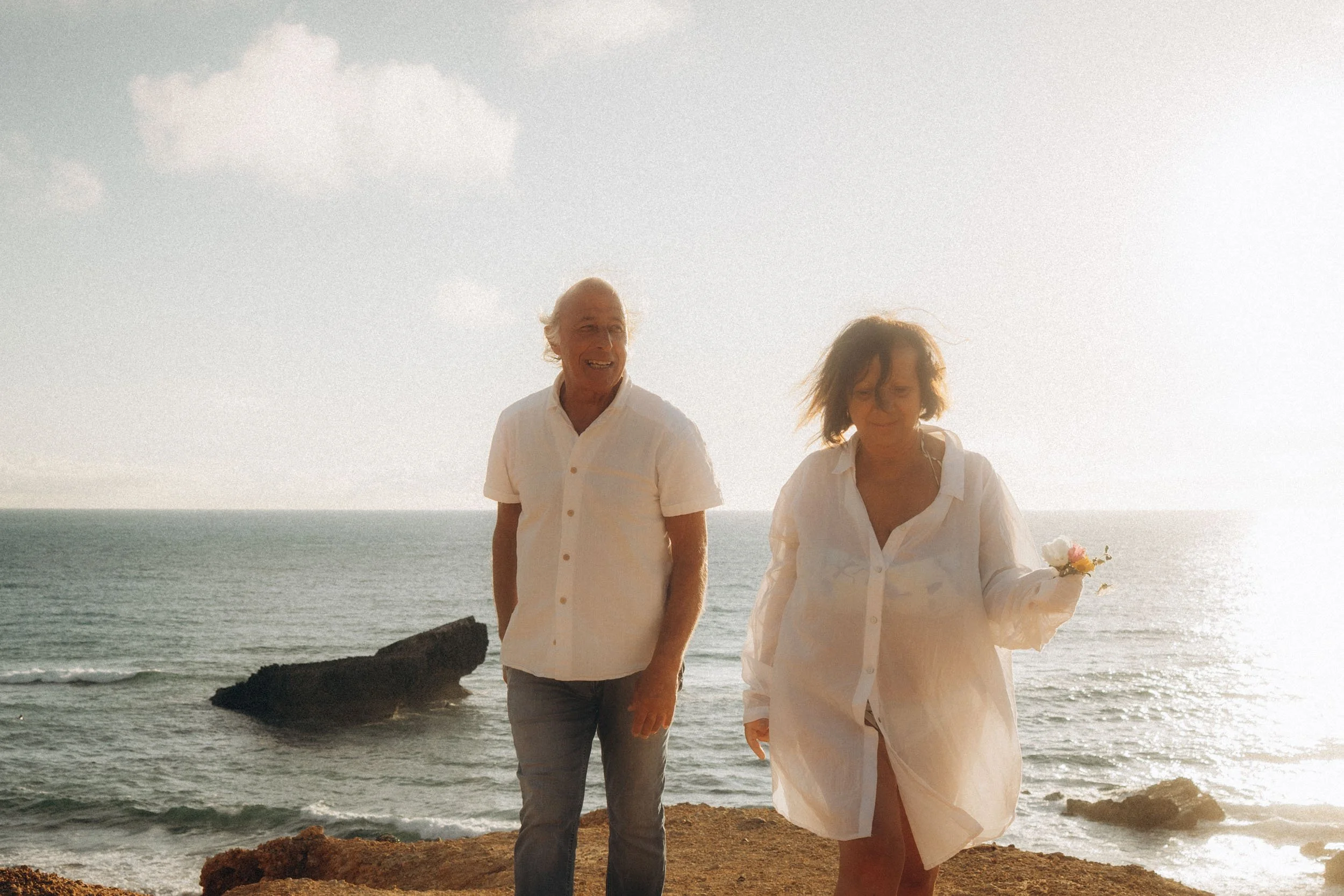 A man and woman walking along a rocky beach at sunset with the ocean in the background.  Algarve couple photographer