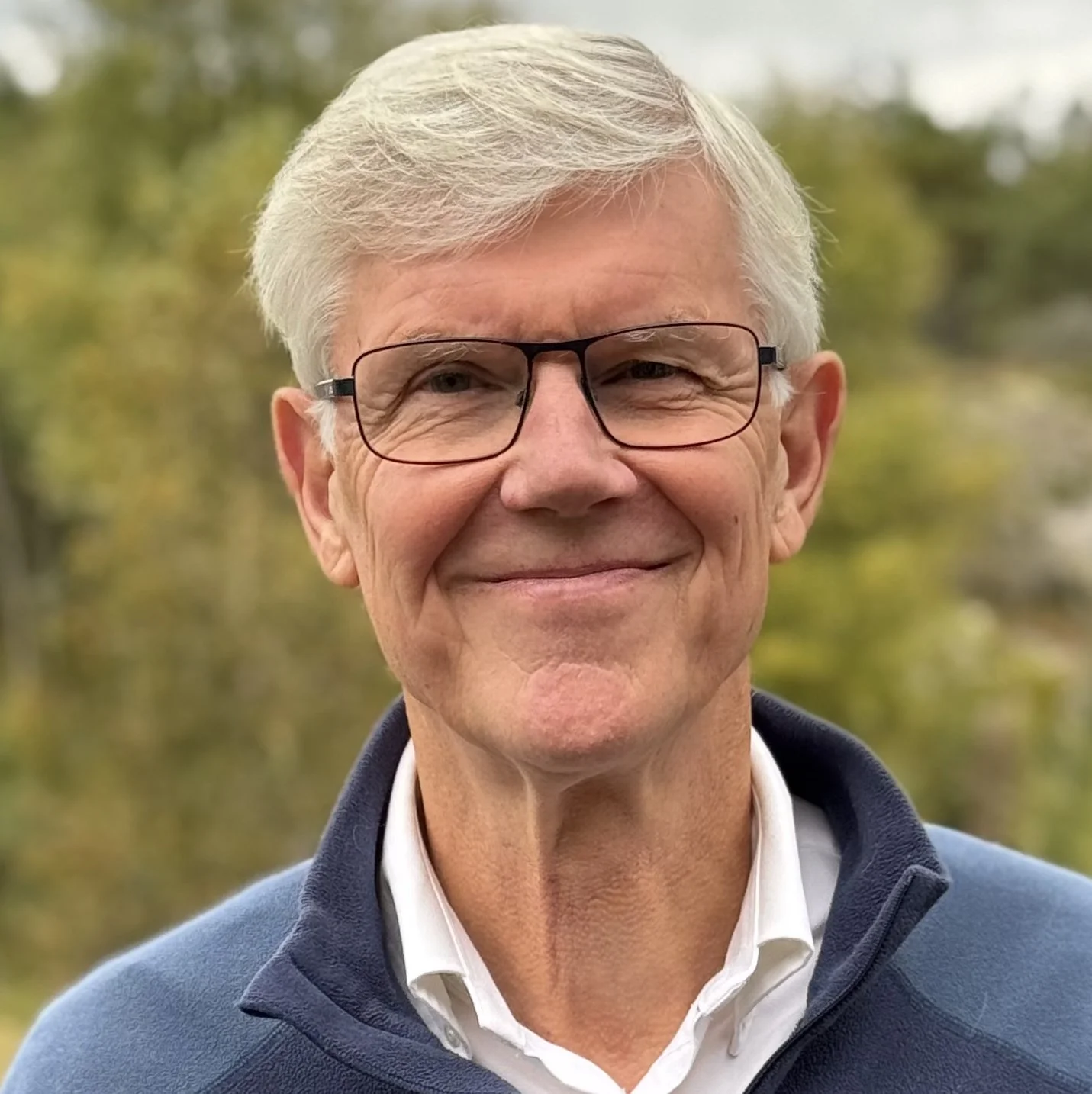 A smiling older man with gray hair, glasses, wearing a white shirt and a blue zip-up jacket, outdoors with blurred green trees in the background.