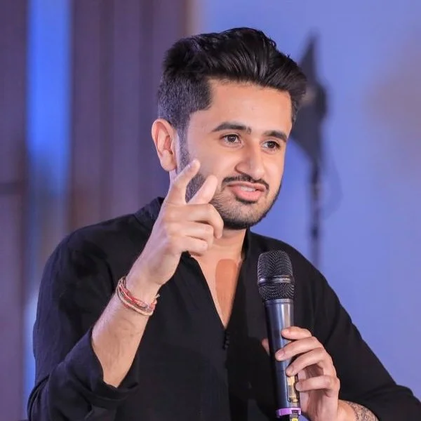 Man speaking into a microphone with a serious expression, pointing with his index finger.