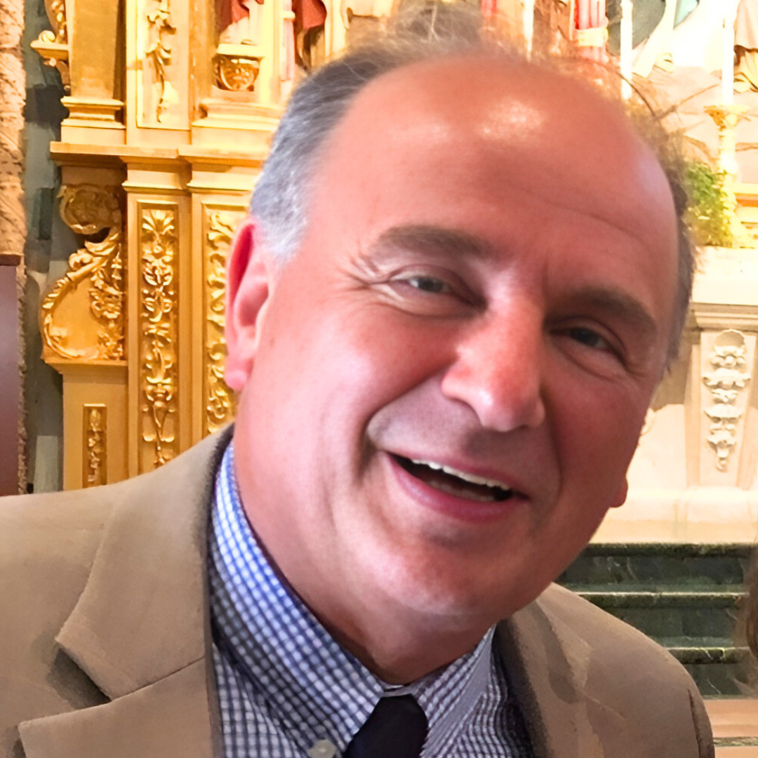 A smiling middle-aged man with short gray hair, wearing a beige blazer, light plaid shirt, and a dark tie, standing inside a building with ornate gold decorations and marble steps in the background.