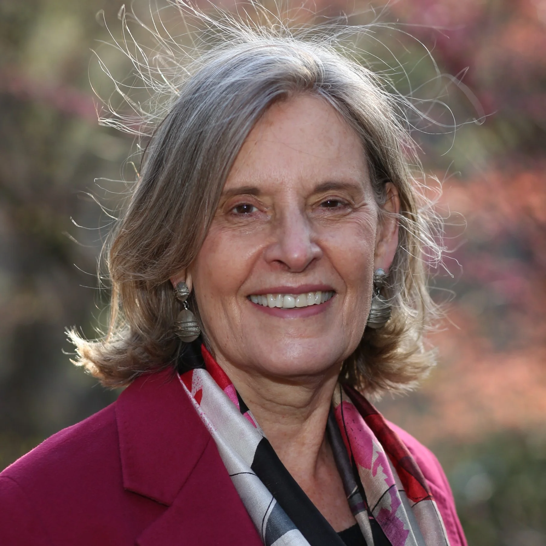 A smiling woman with shoulder-length gray hair, wearing a magenta blazer and a colorful scarf, standing outdoors with blurred trees in the background.