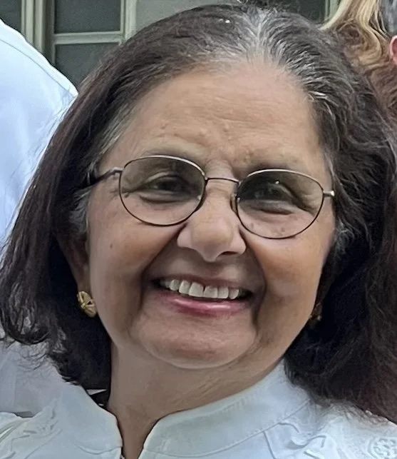 A smiling middle-aged woman wearing glasses and gold earrings, with shoulder-length dark hair, outdoors.