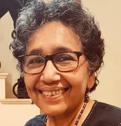 Close-up photo of a smiling woman with curly gray hair, glasses, and earrings.