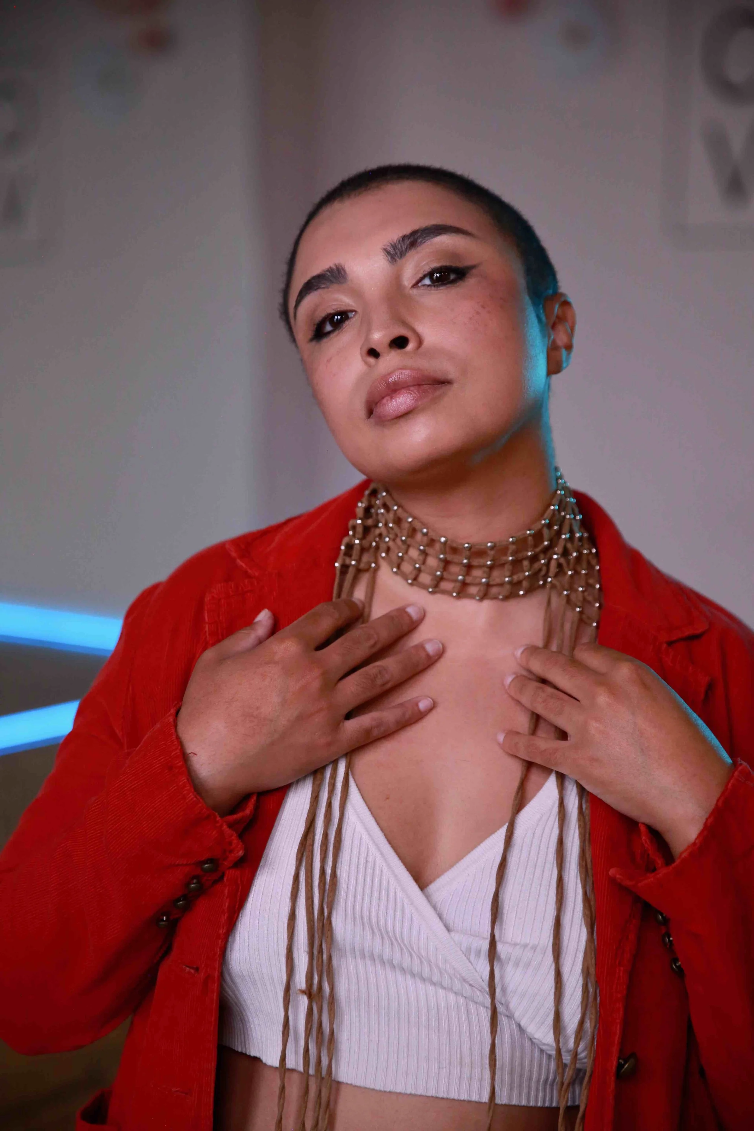 A woman with short hair and makeup, wearing a red jacket over a white top, accessorized with layered necklaces, looking confidently at the camera.