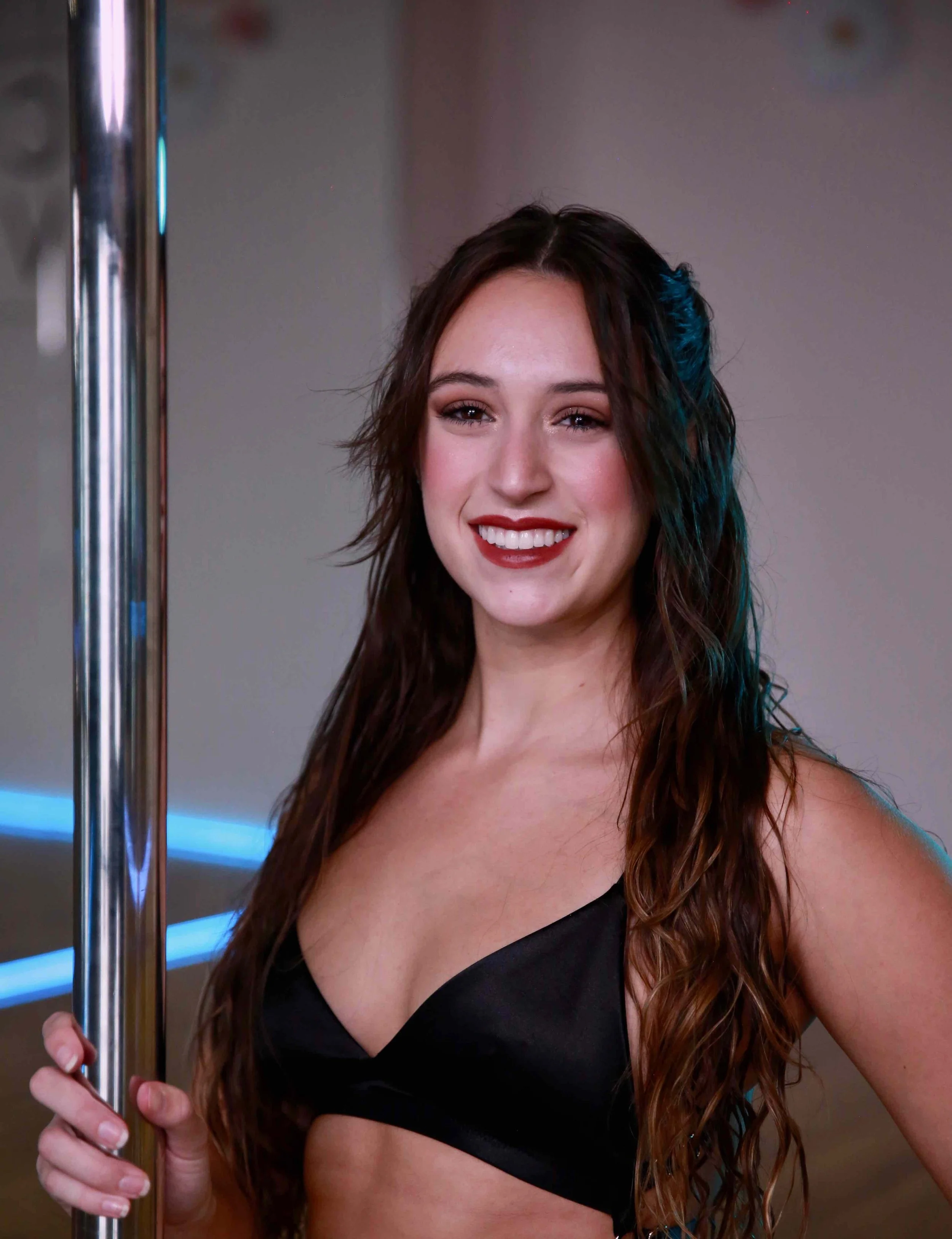 Young woman with long wavy brown hair wearing a black dance bra, holding a metal pole, smiling in a dance studio with blue lighting.