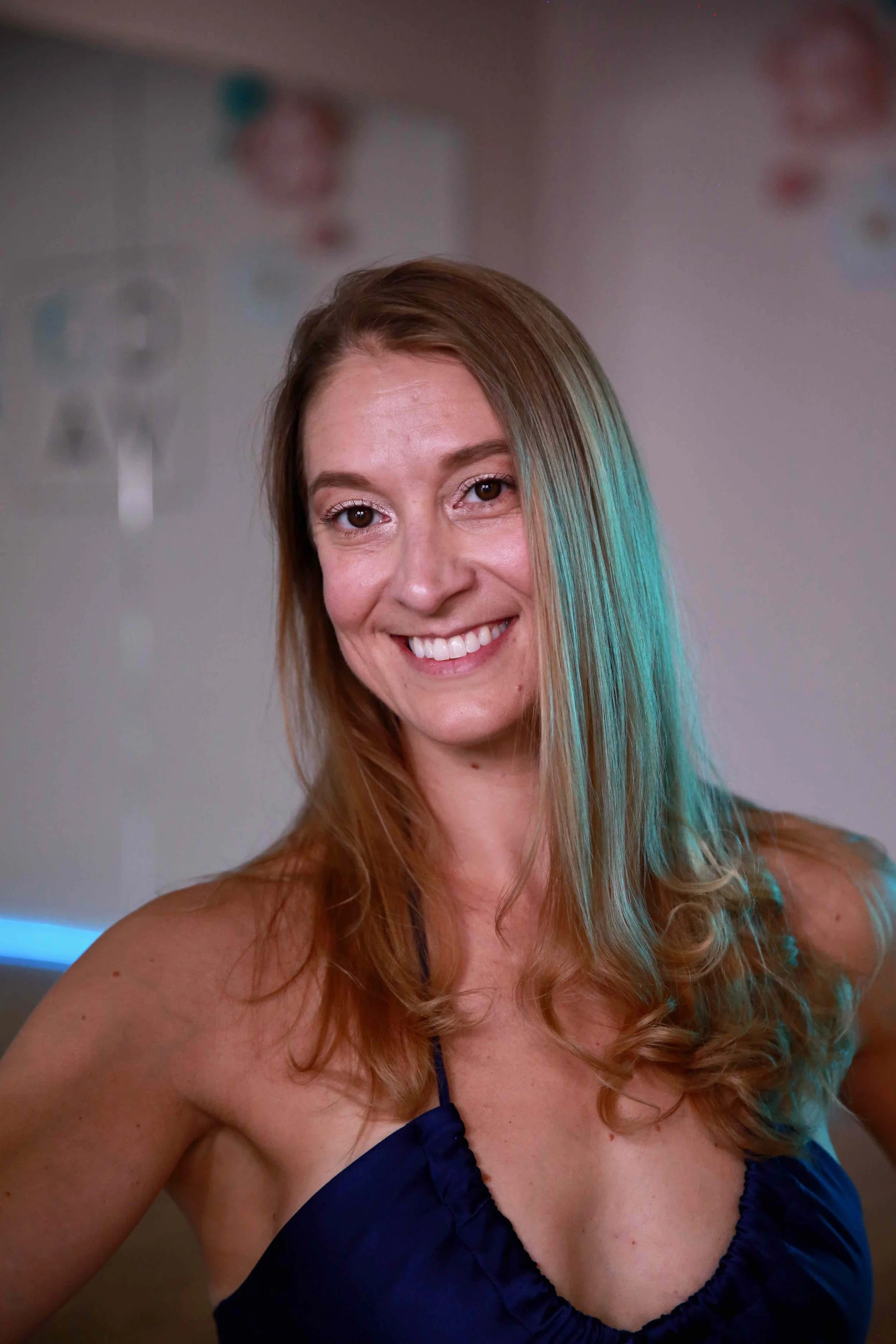 A woman with long, wavy brown hair smiling at the camera in a room with a blurred background.