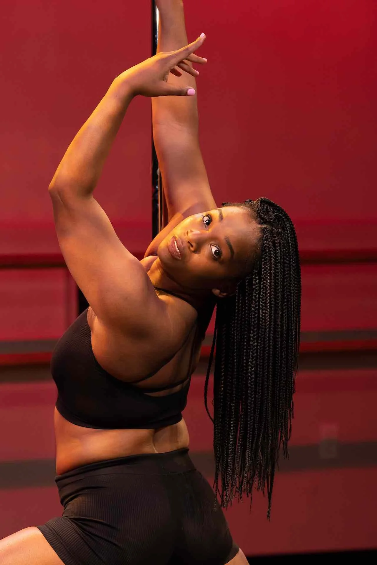 A woman with long braided hair performs a dance move against a red background, wearing a black sports bra and black shorts.