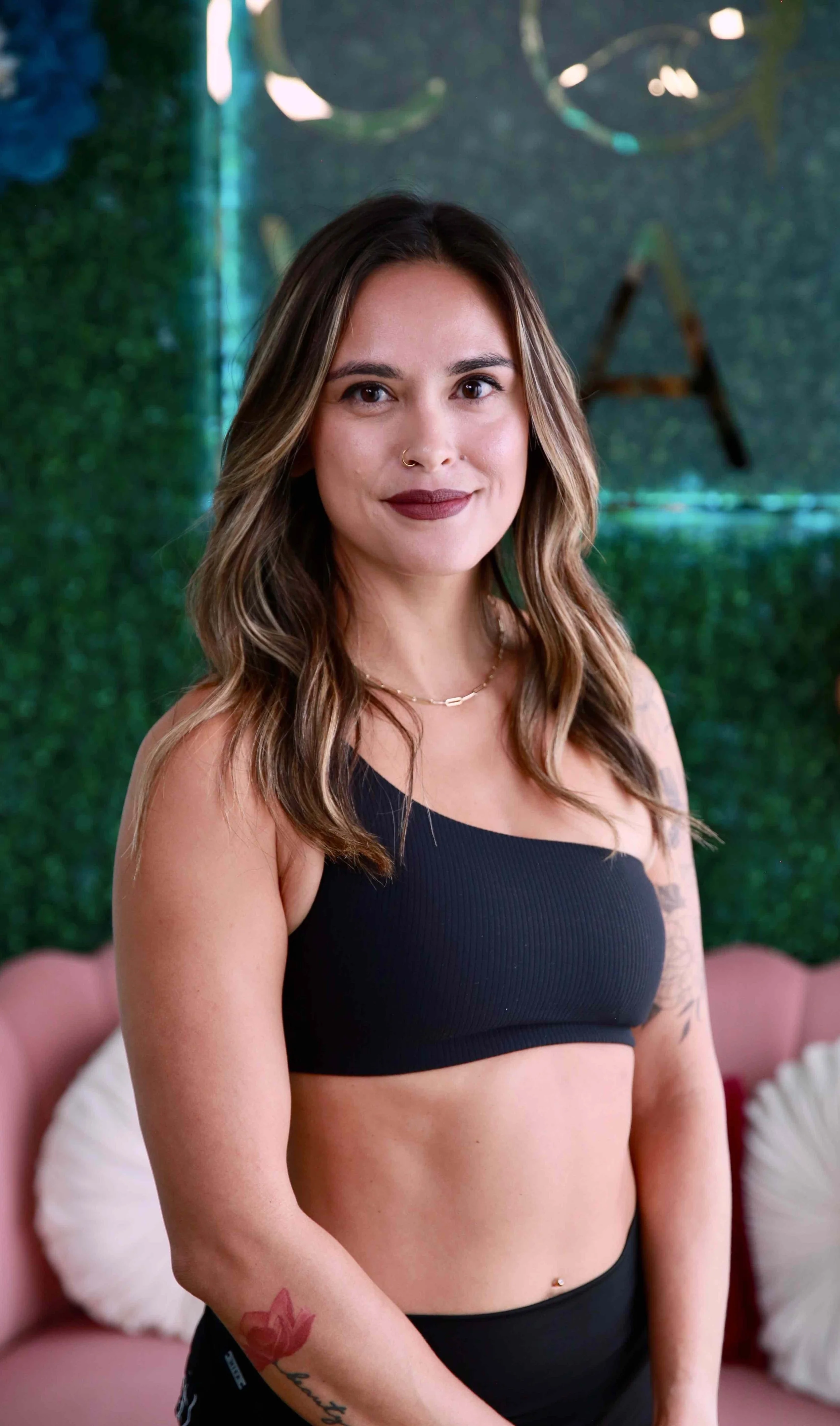 Young woman with long, wavy brown hair wearing a black sports bra and black shorts, standing in front of a green textured wall with decorative lighting.