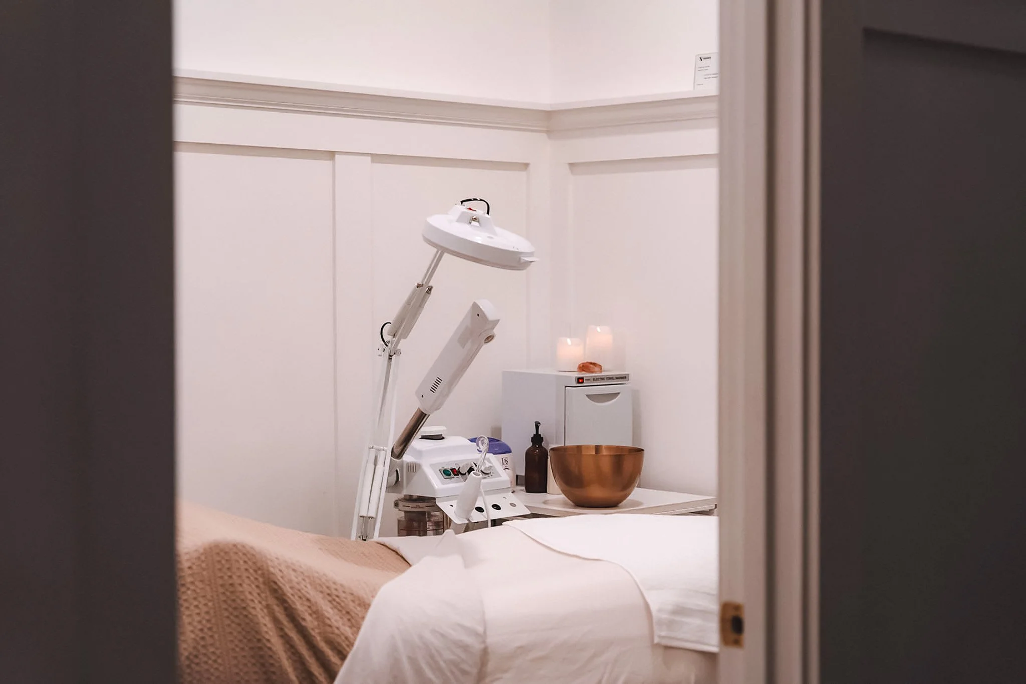 A medical examination room seen through door frame, with a bed, a brown bowl, a white medical lamp, and various medical devices and paraphernalia.
