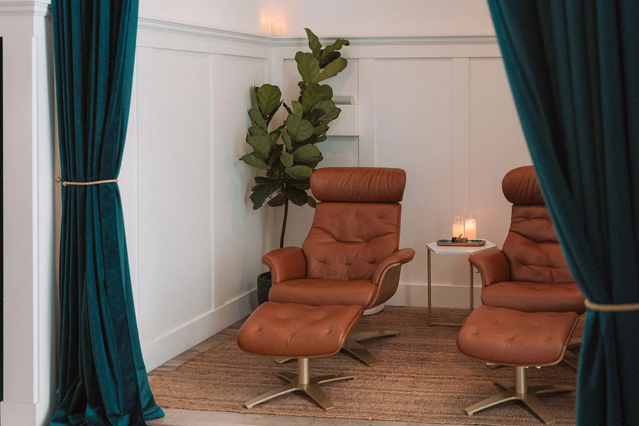 A cozy room with two brown leather lounge chairs, a side table with a lit candle and decorative items, a potted fiddle leaf fig plant, white paneled walls, teal curtains pulled back, and soft lighting.