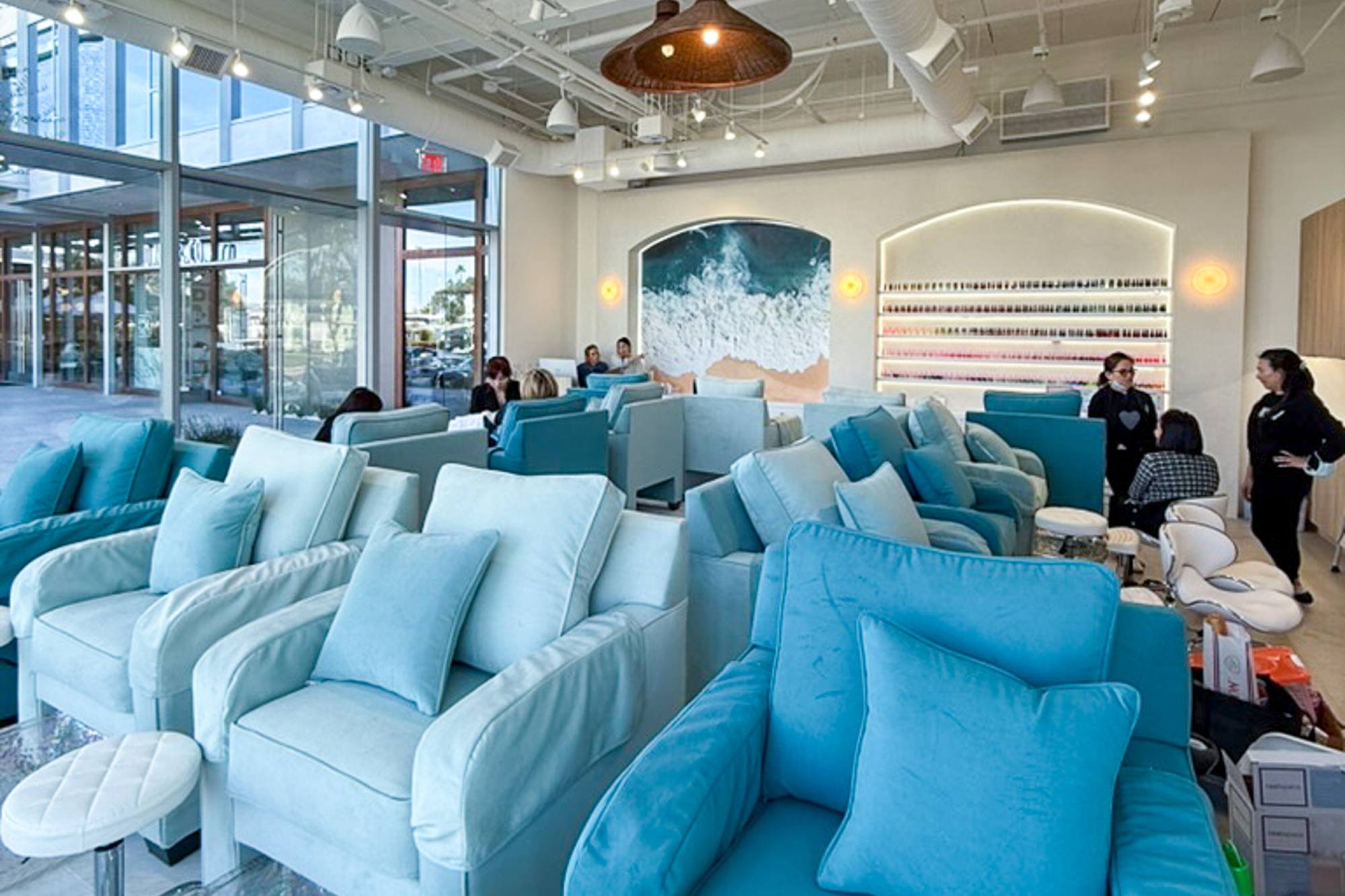 Exterior view of a modern nail salon with large glass windows, showcasing an interior with teal seating and nail stations at night, surrounded by landscaping.