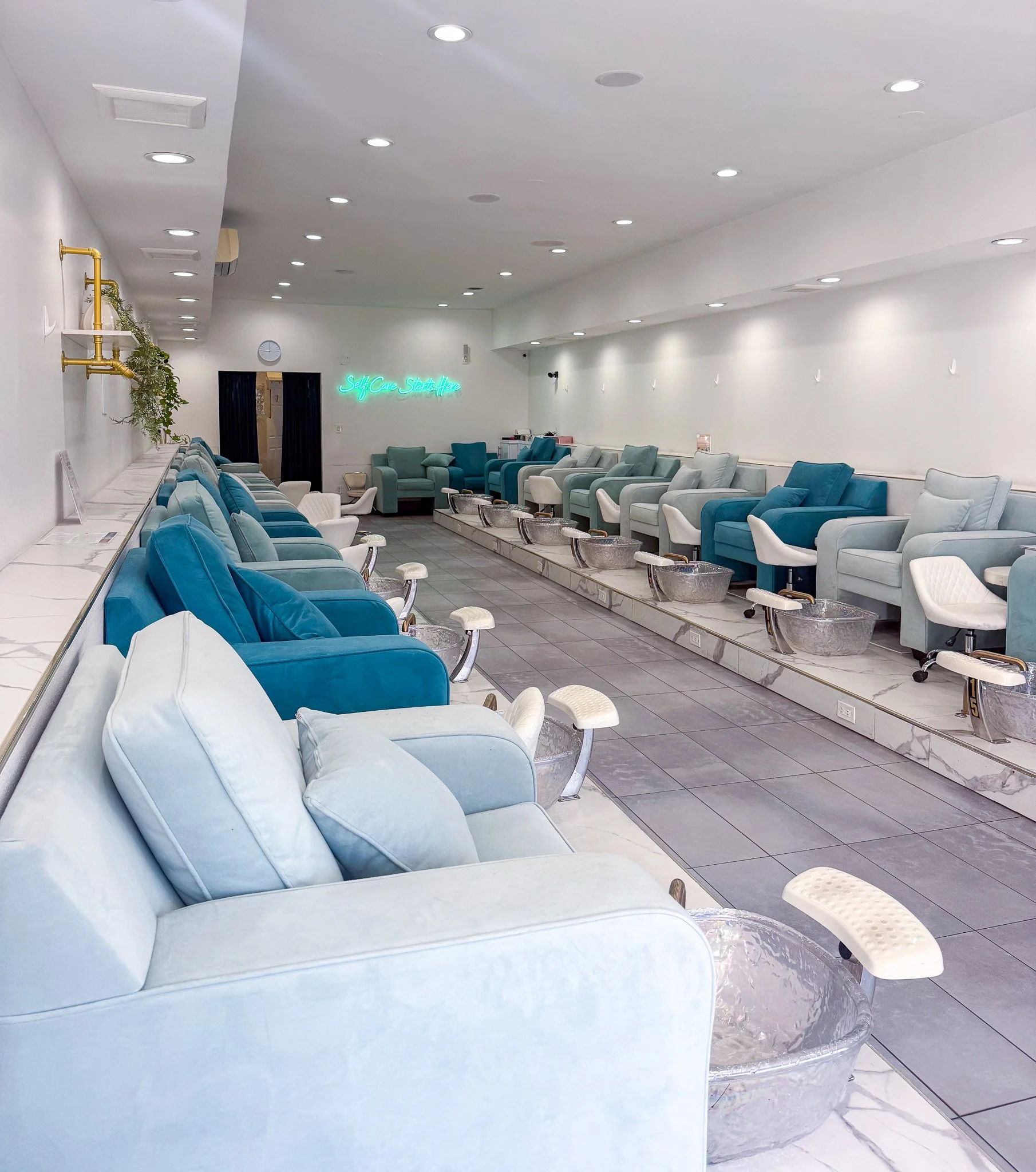 Nail salon with white walls, ceiling of recessed lights, and chairs with foot baths, some chairs have teal and white upholstery, a neon sign that says 'Self Care Starts Here', and a clock on the wall.