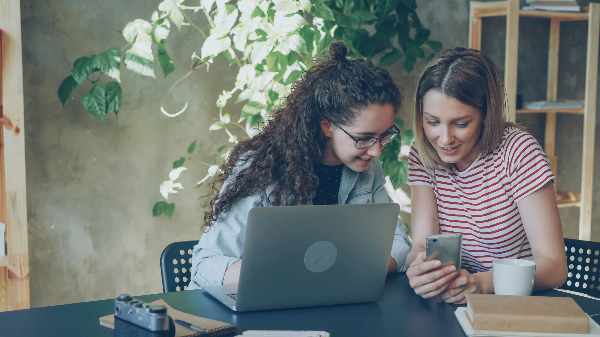 Two young women sitting at a table, looking at a smartphone together, with a laptop, camera, notebooks, a mug, and a book on the table, in a room with green plants and wooden shelves.