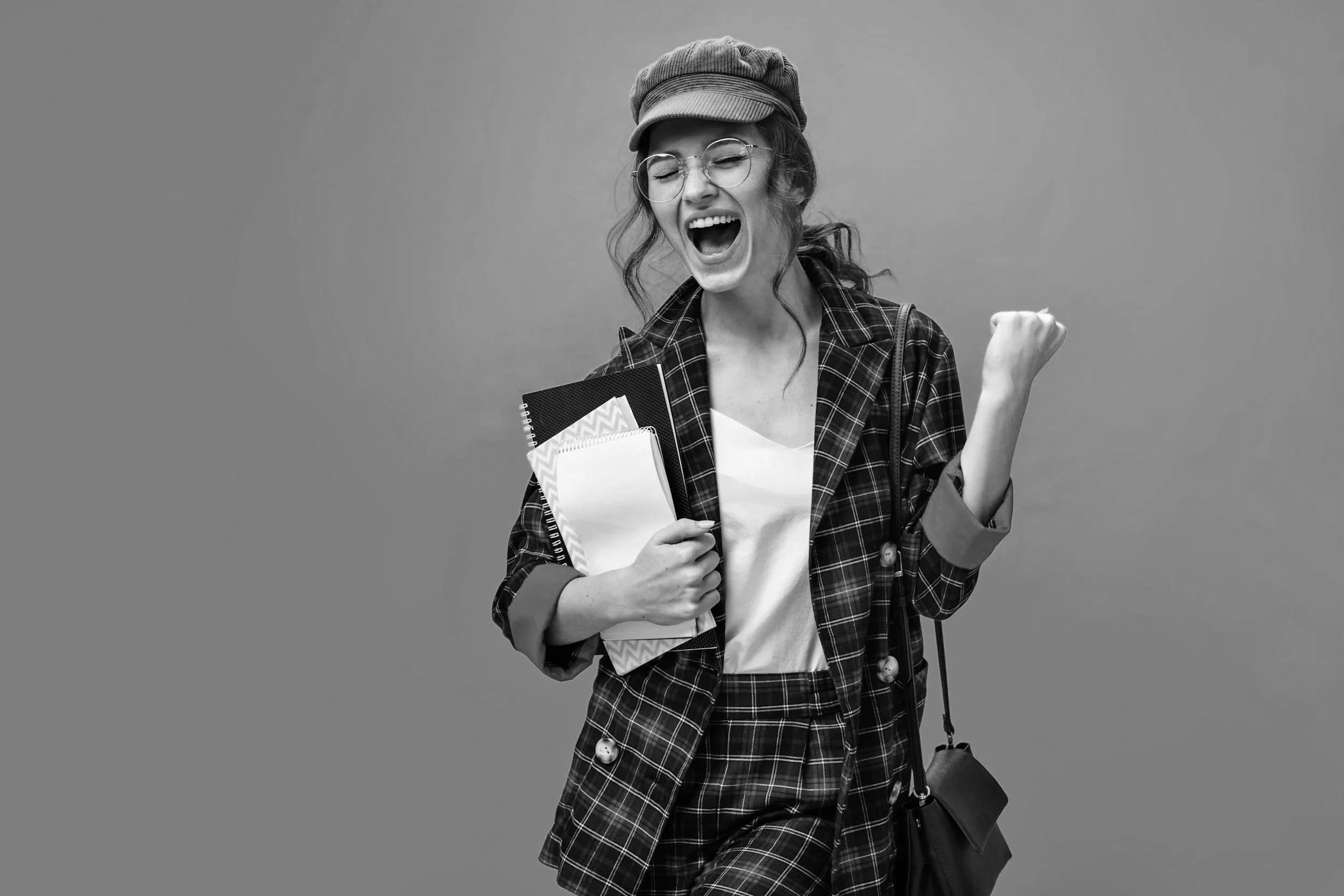 A young woman wearing glasses, a hat, and a plaid jacket, holding notebooks and smiling with a fist pump gesture against a plain background.