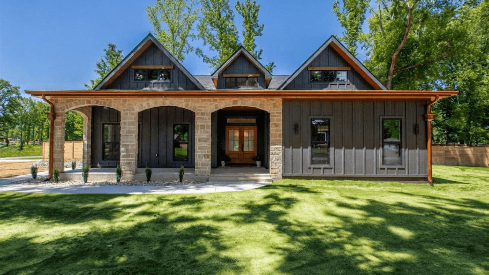 Front view of a modern two-story house with stone and dark wood exterior, large front yard, and green trees in the background.