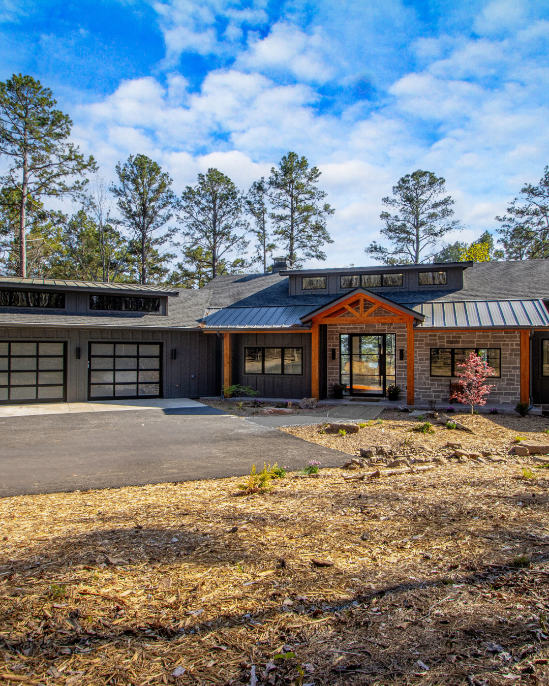 Modern single-story house with a dark gray exterior, large windows, and a wooden porch entrance, surrounded by trees and a partly cloudy sky.