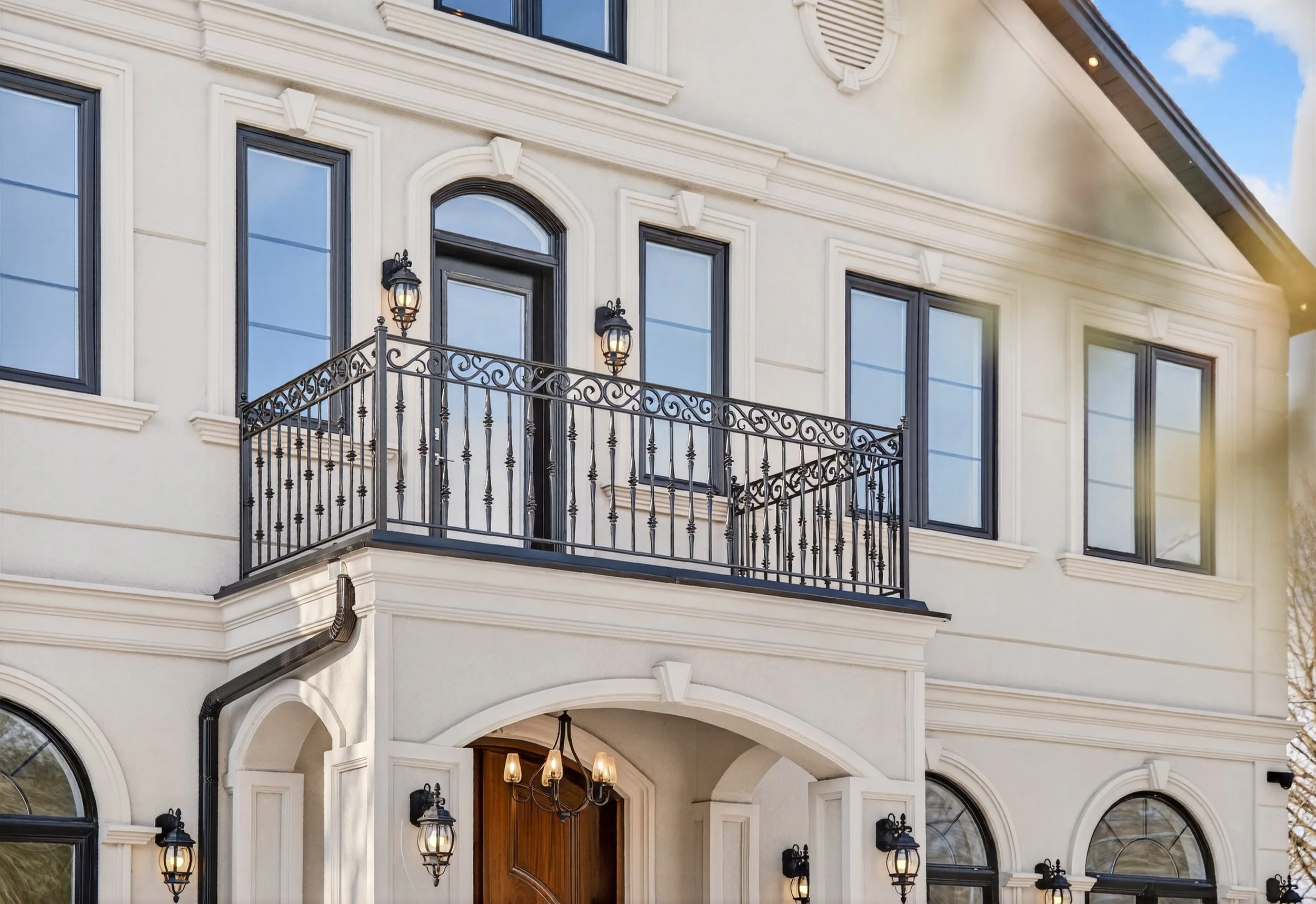 The image shows the facade of a luxurious, multi-story house with cream-colored walls and large black-framed windows. There is a small balcony with decorative black wrought-iron railings and two wall-mounted lantern lights above the balcony door. The entrance is adorned with modern black lanterns and a wooden door beneath a stone archway. The sky is partly cloudy with sunlight illuminating part of the house.