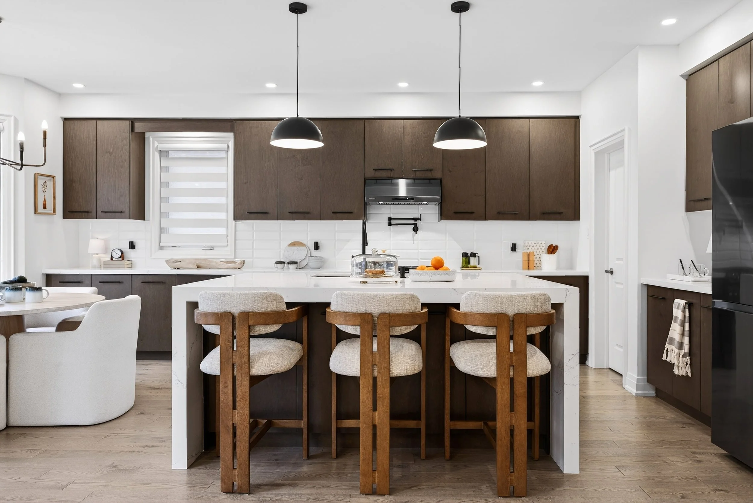 Modern kitchen with dark wooden cabinets, white countertop, and a central island with seating. Black pendant lights hanging above the island, a window with a white blind, and various kitchen utensils and decor.