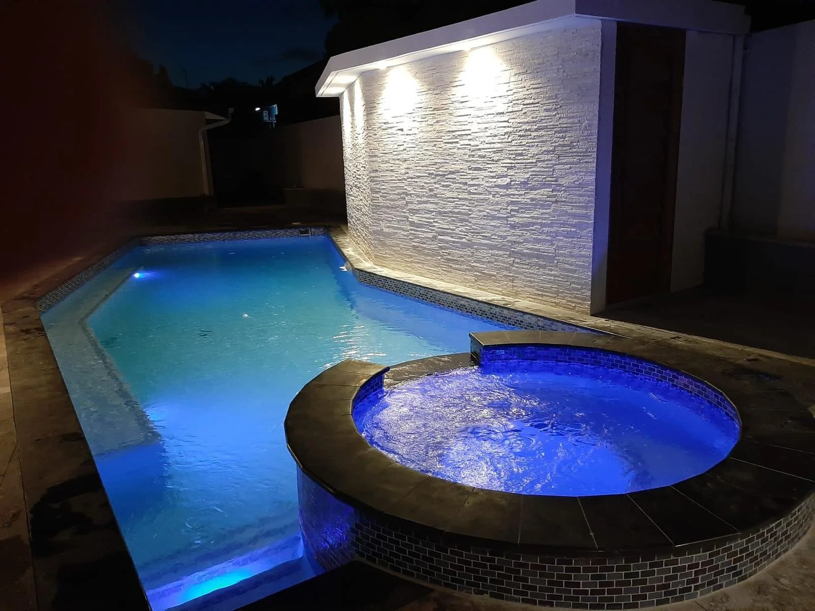 Een verlichte privézwembad en jacuzzi 's avonds, met blauwe verlichting en een wit siermuur.
