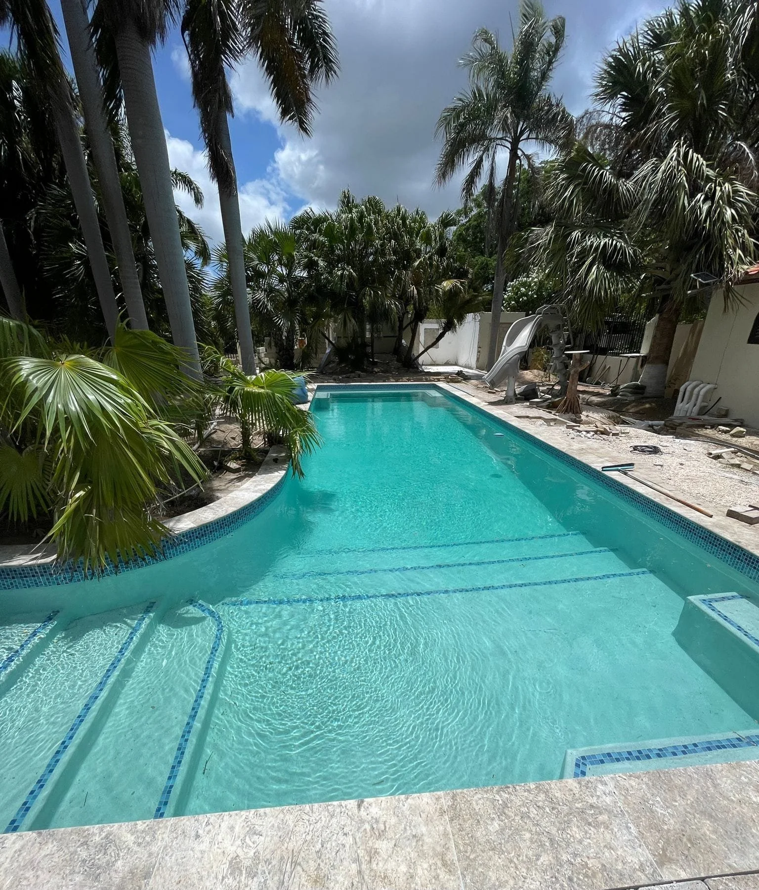 Een zwembad met blauwe water, omgeven door palm- en andere tropische planten, in een tuin met een gedeeltelijk beschadigde rand en een glijbaan op de achtergrond, onder een bewolkte lucht.