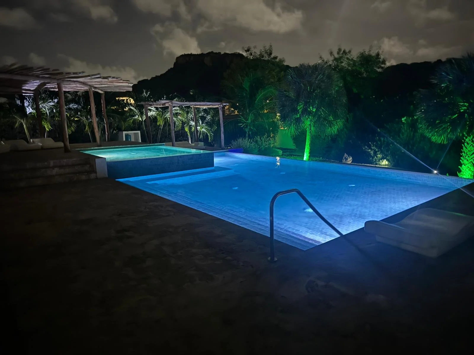 Zwembad bij nacht met verlichte omgeving, palmbomen en een berg op de achtergrond, verlichting in verschillende kleuren