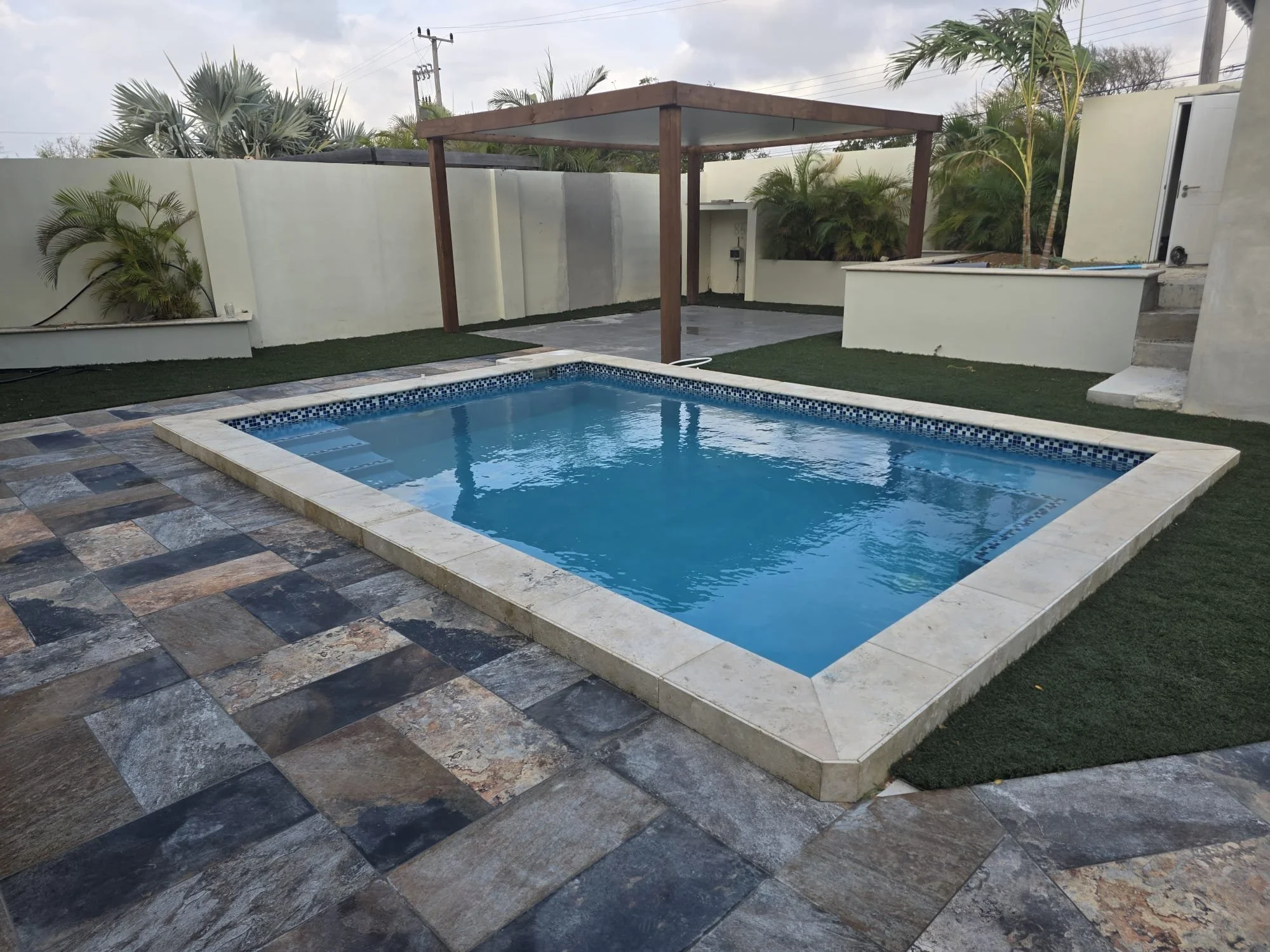 Een tuin met een klein zwembad, betegeld met multicolor tegels, omgeven door kunstgras en een witte muur, met een houten pergola en groene planten op de achtergrond.