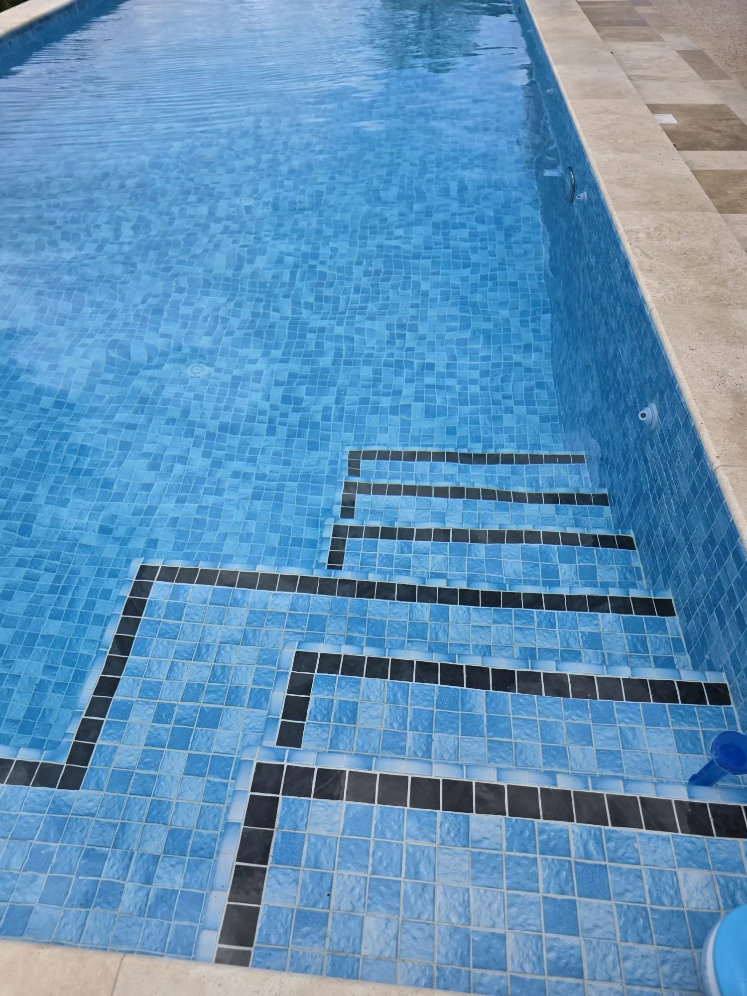 De afbeelding toont een zwembad met blauwe water en blauwe tegels, omringd door betegelde vloer, met zwarte tegelpatronen op de rand van het zwembad.