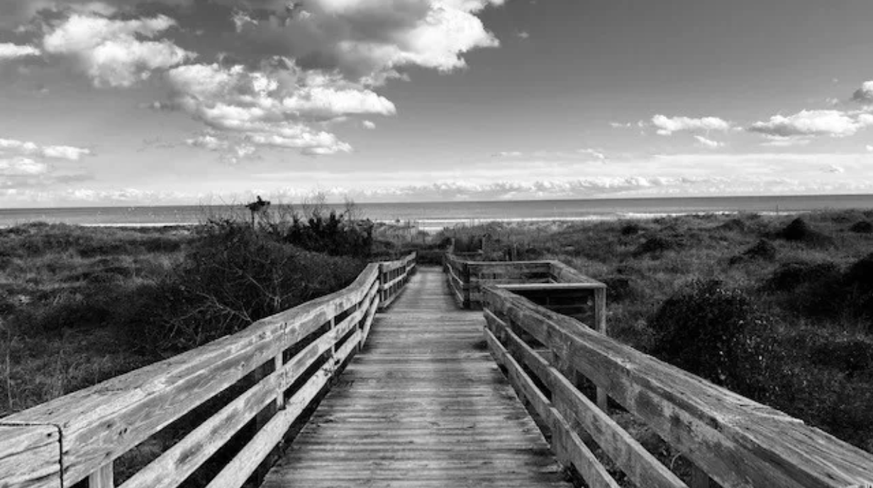 A wooden boardwalk leading towards the beach with sand dunes and bushes on either side, under a partly cloudy sky.